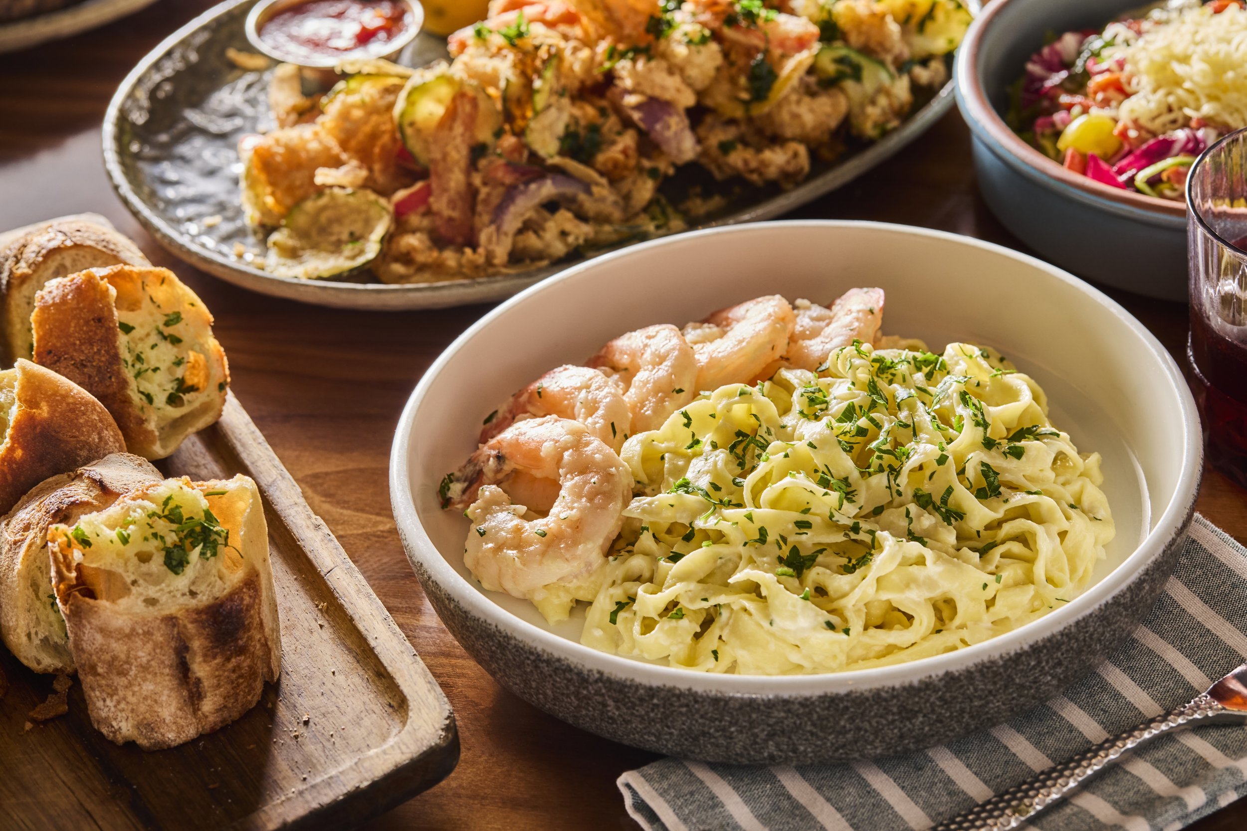 Assorted dishes of pasta, salad, bread rolls, and fried appetizers on a wooden table with a striped napkin.