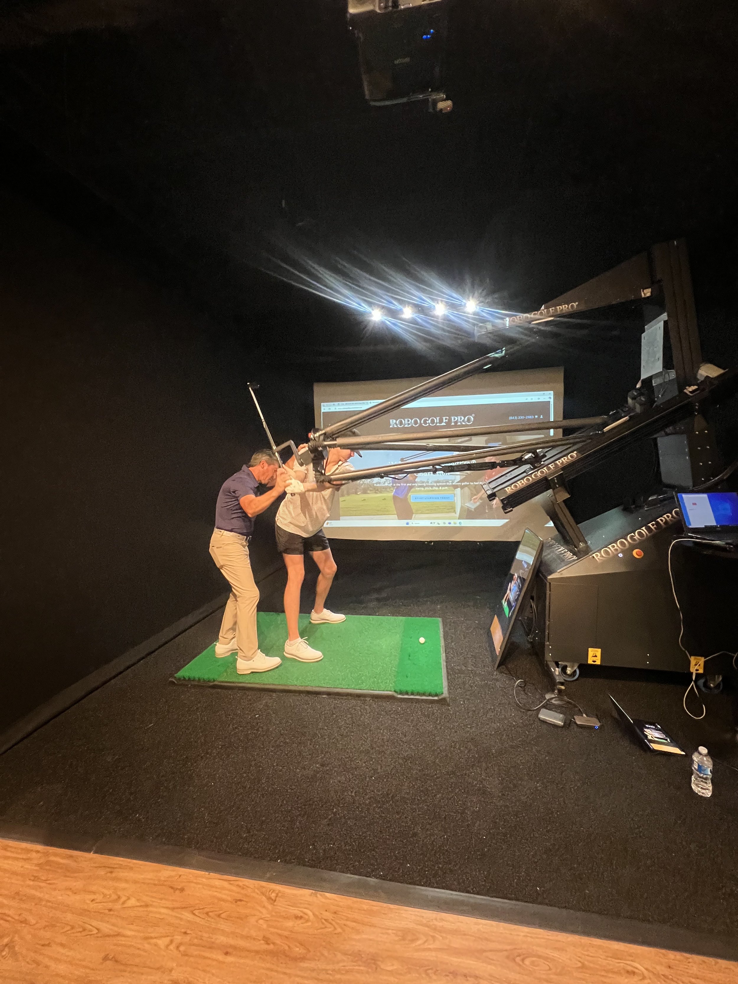Two people in a golf simulator, with a RoboGolfPro machine, practicing swings on a green mat in an indoor facility.