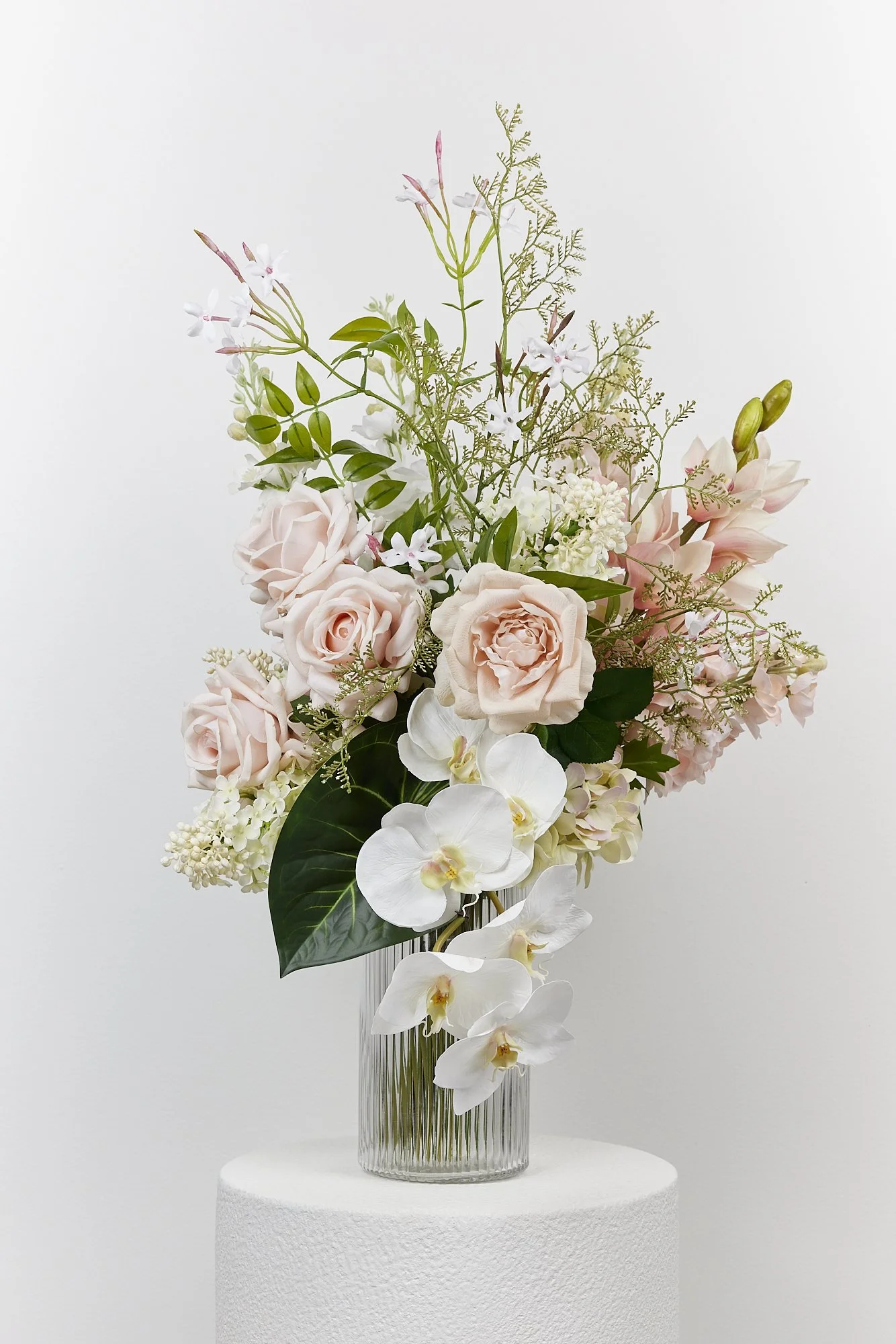 A tall glass vase with white and blush pink flowers, including roses, orchids, and other assorted blooms, arranged with green foliage, set on a white textured pedestal against a plain white background.