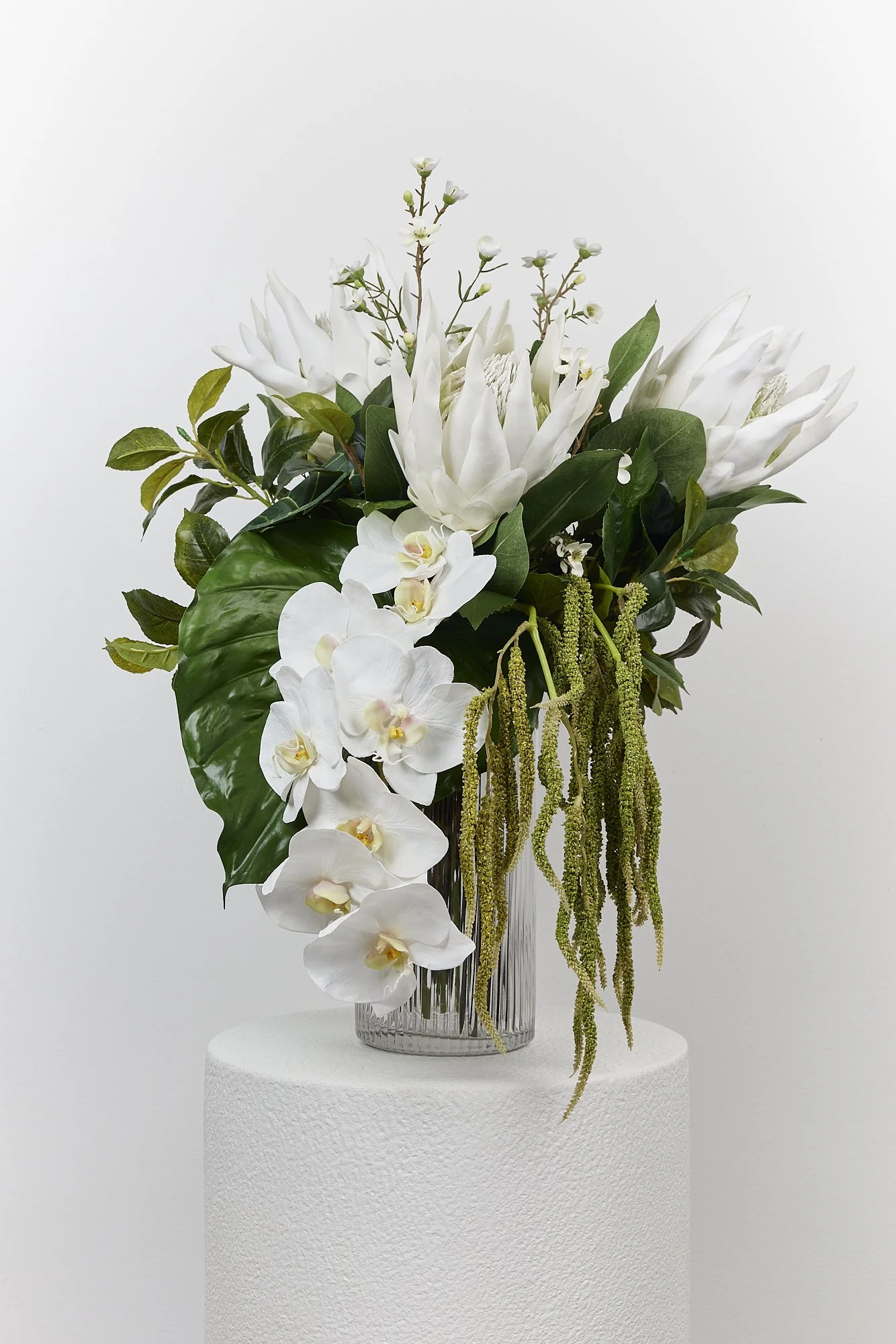 White flower arrangement with orchids, magnolia blossoms, and green foliage in a glass vase on a white pedestal against a white background.