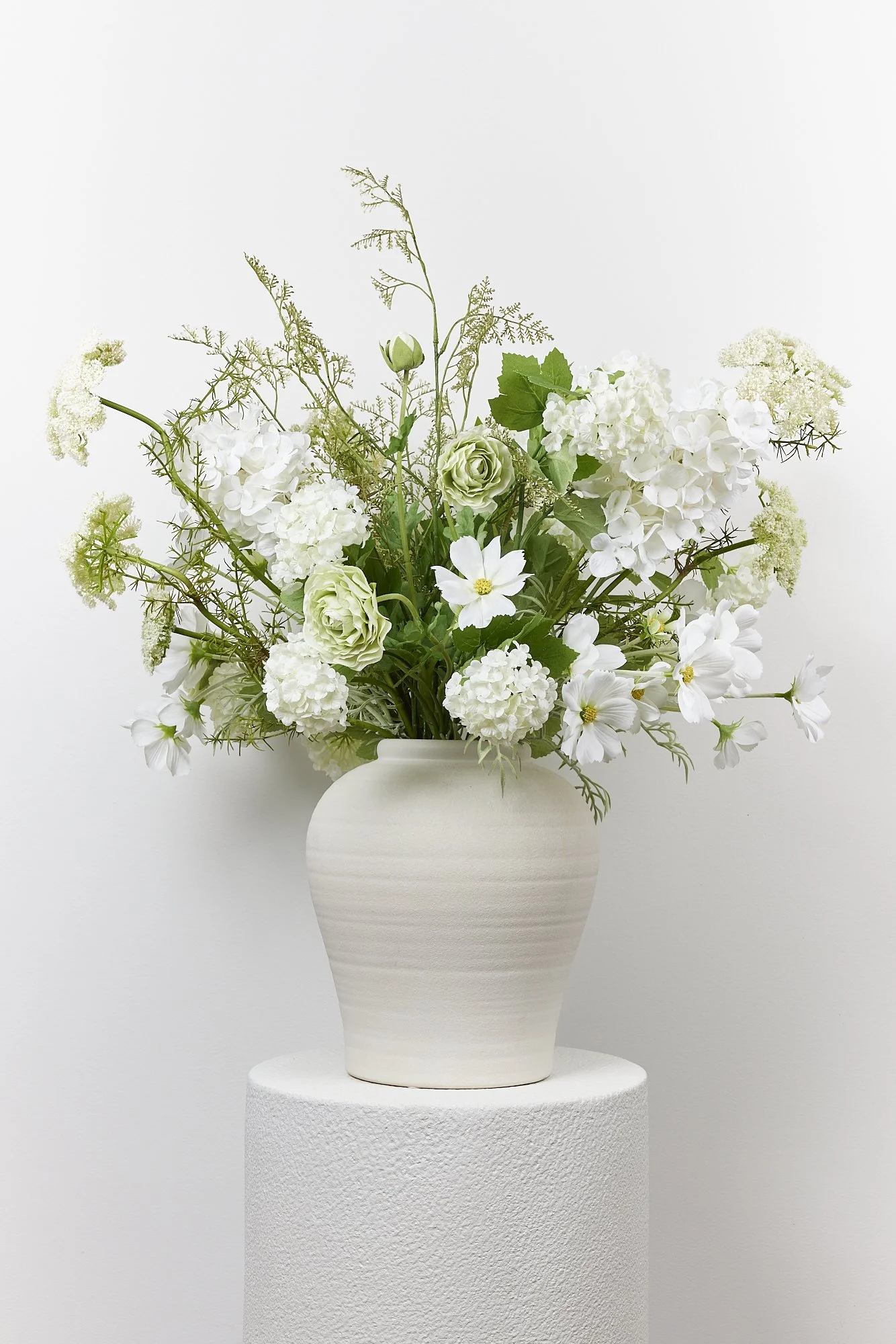 White ceramic vase with a bouquet of white flowers, including hydrangeas, daisies, and other blooms, on a white textured pedestal against a plain white background.
