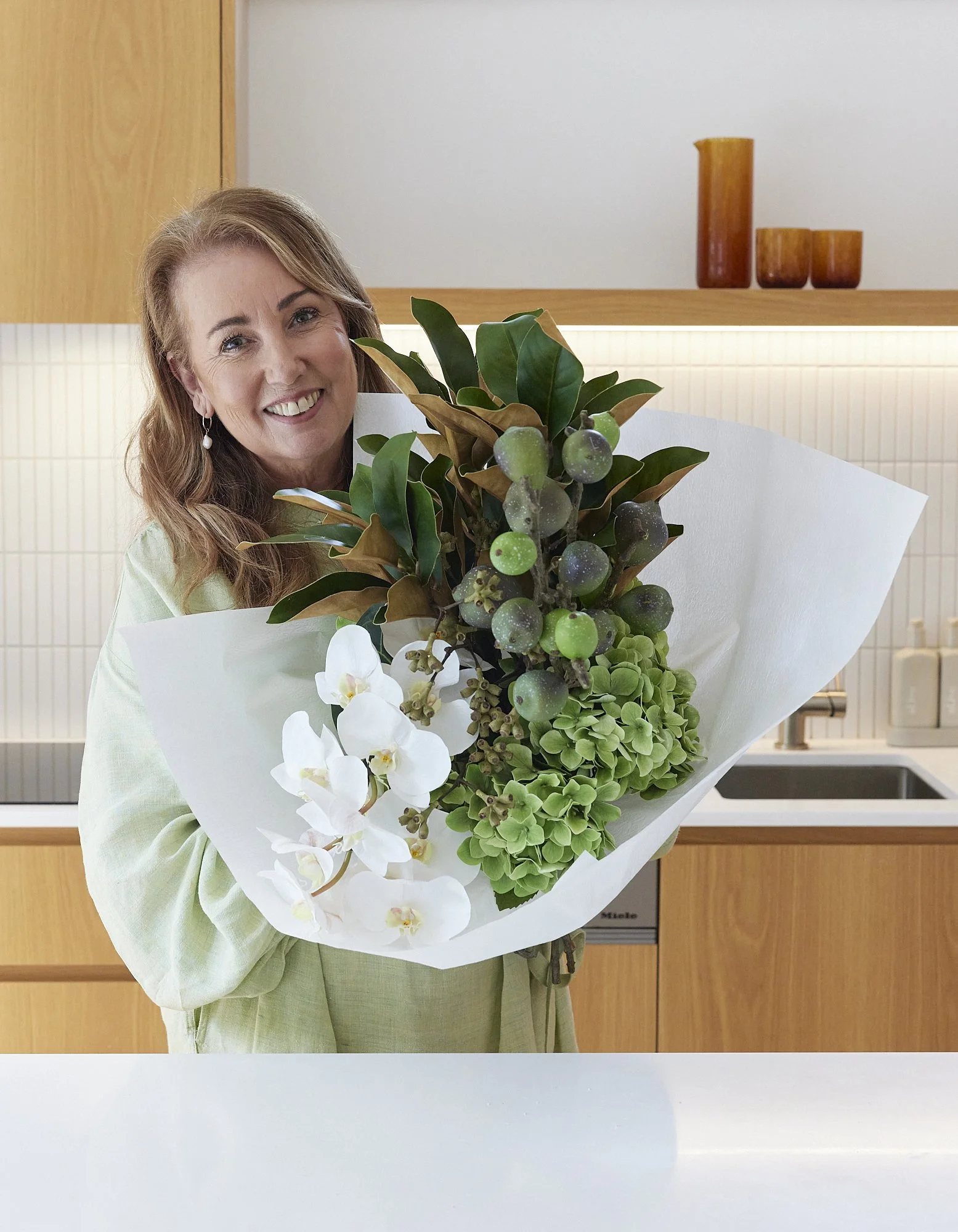 A woman holding a large bouquet of white orchids, green hydrangeas, and foliage in a modern kitchen.