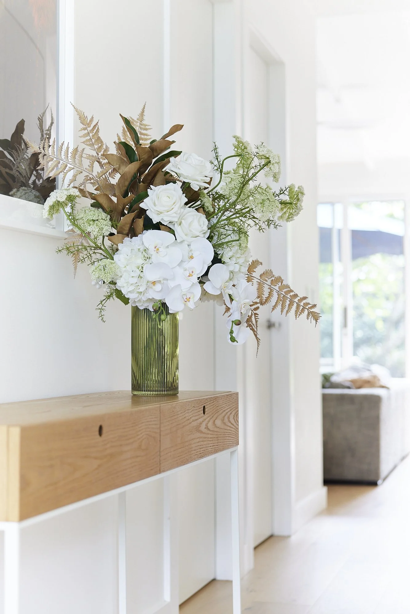 A floral arrangement with white roses, orchids, and greenery in a textured green vase on a wooden console table in a bright, modern interior.