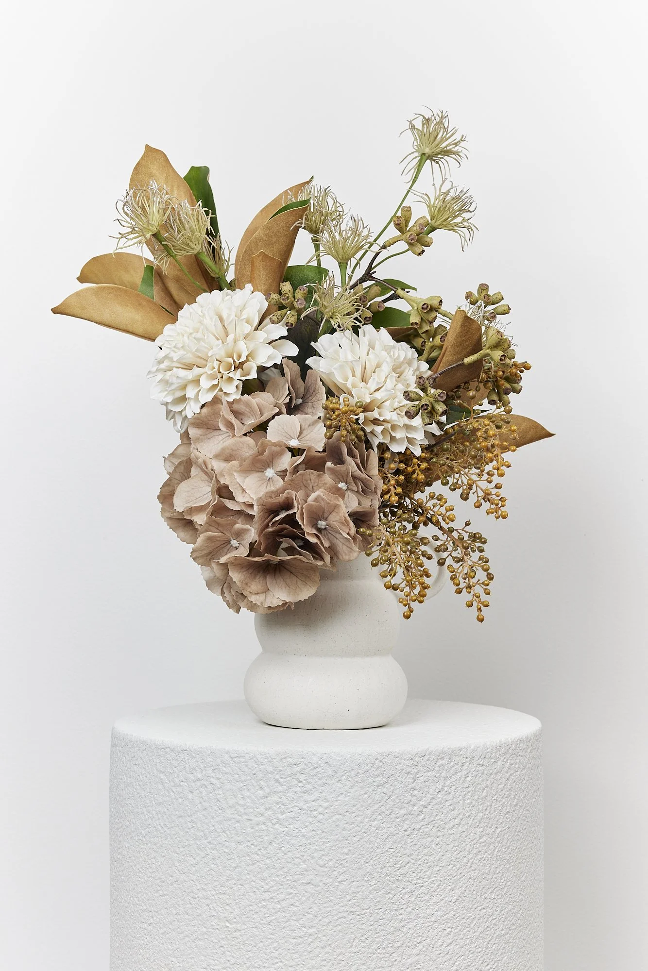 A white ceramic vase with a textured surface holding a bouquet of dried beige, white, and green flowers and foliage, placed on a white textured cylindrical pedestal against a plain white background.