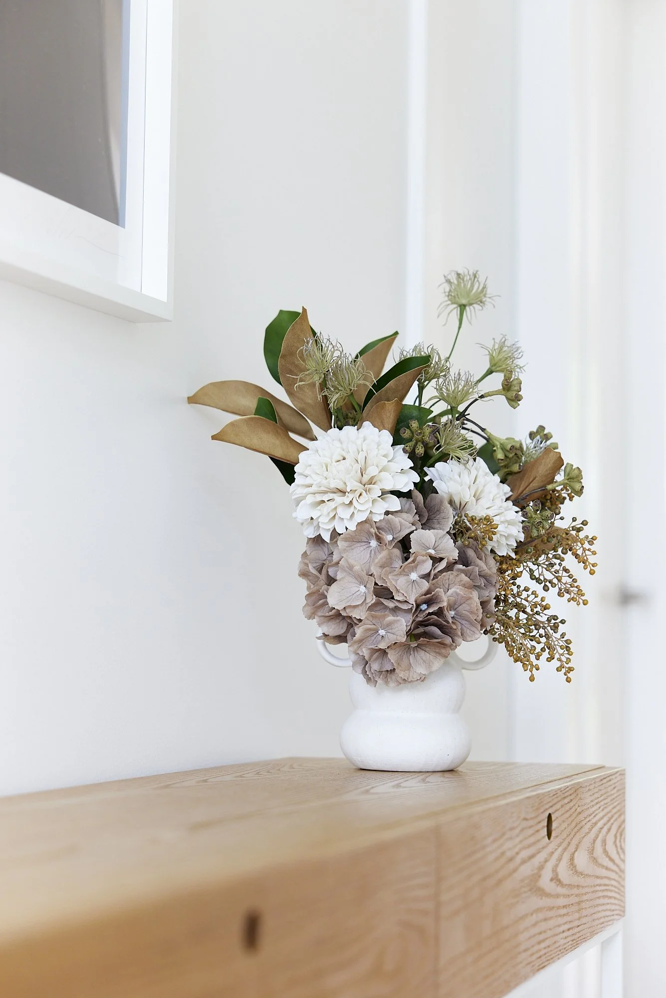 A white vase with a floral arrangement on a wooden surface, containing white, beige, and green flowers and leaves, against a plain light background.