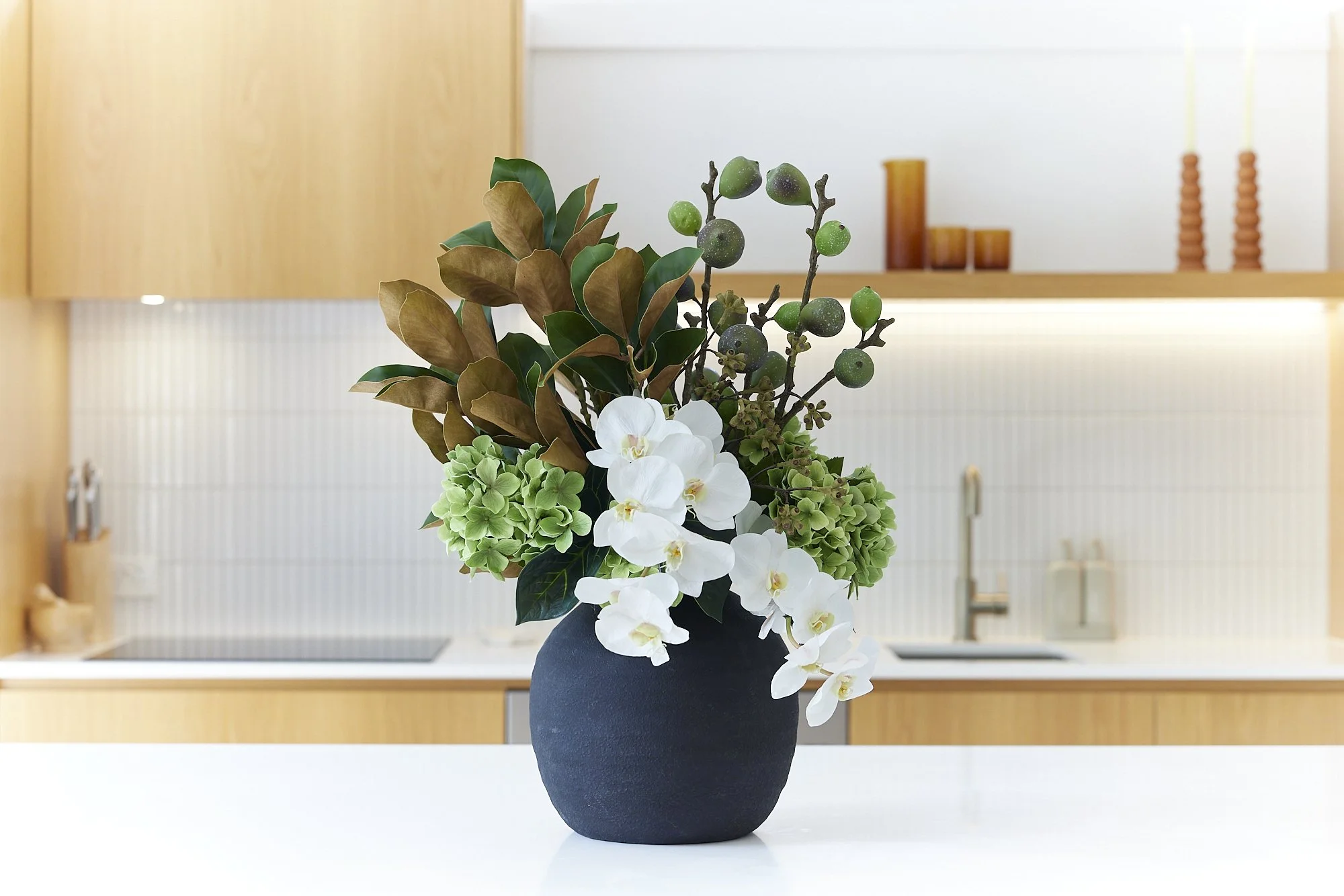 A black vase with white orchids, green hydrangeas, brown and green leaves, and green berries on a white countertop in a kitchen with light wood cabinets and white backsplash.