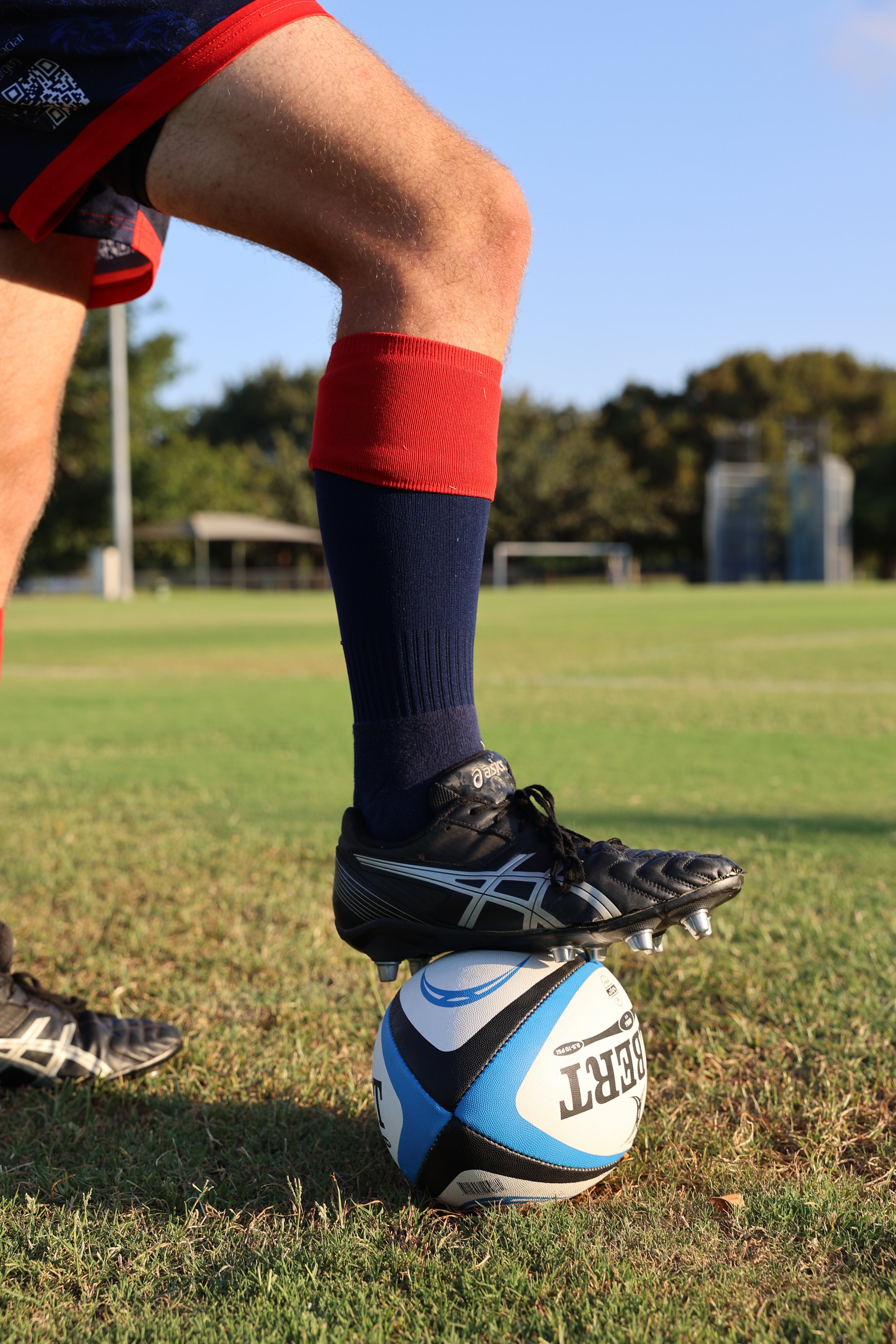 A person standing on a soccer field balancing a soccer ball with their foot, wearing black cleats, navy socks with red and navy sleeves, and red shorts.