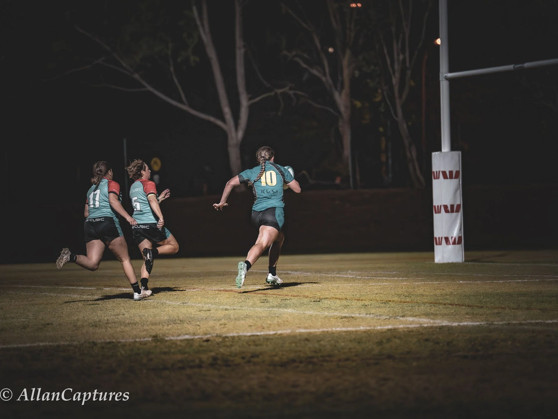 Nighttime rugby match with four female players running towards the goal post on a grass field.