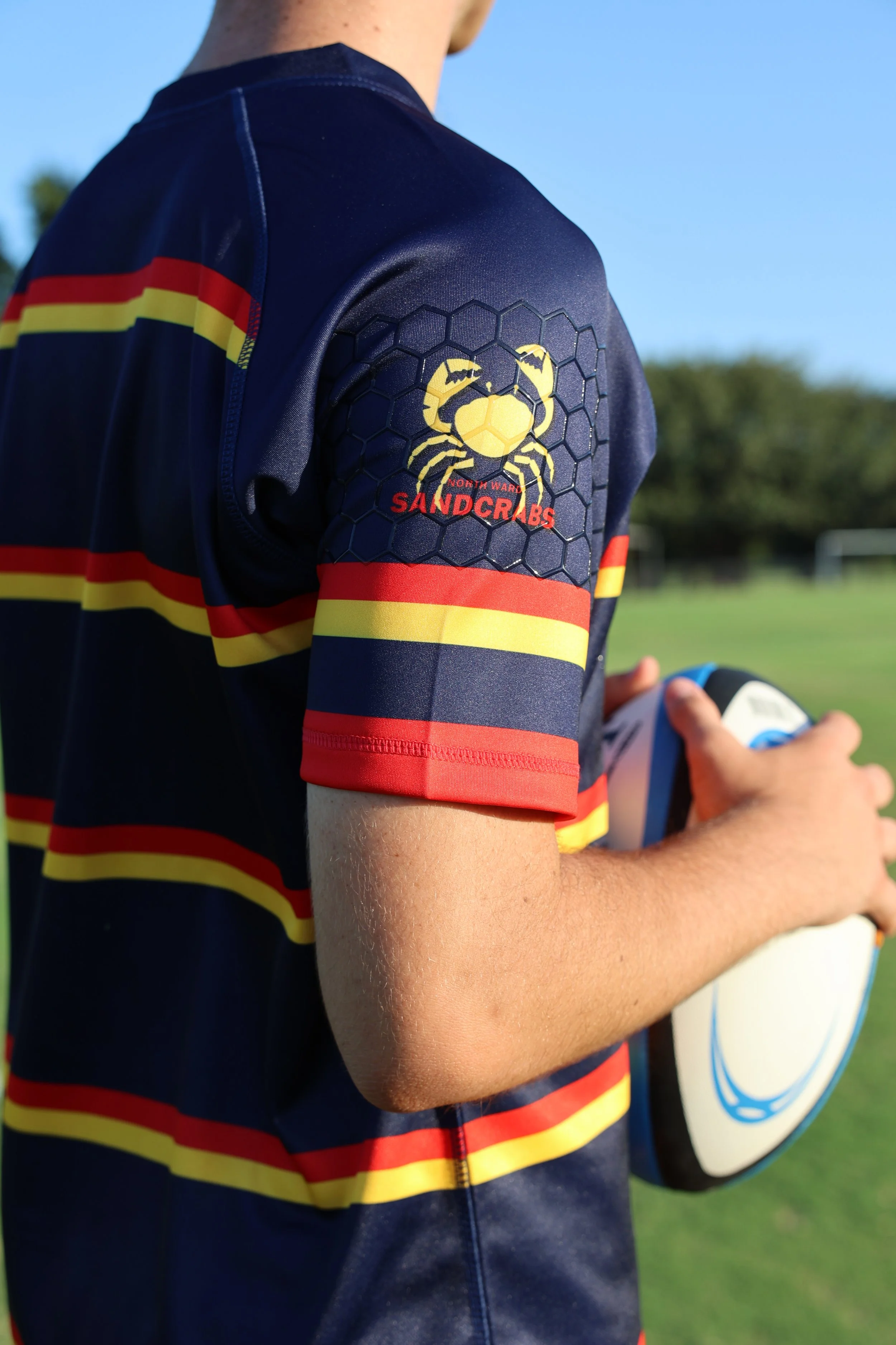Close-up of a rugby player wearing a navy jersey with red, yellow, and navy stripes, holding a rugby ball on a field. The jersey has a logo with a yellow crab and the word 'Sandcrabs'.