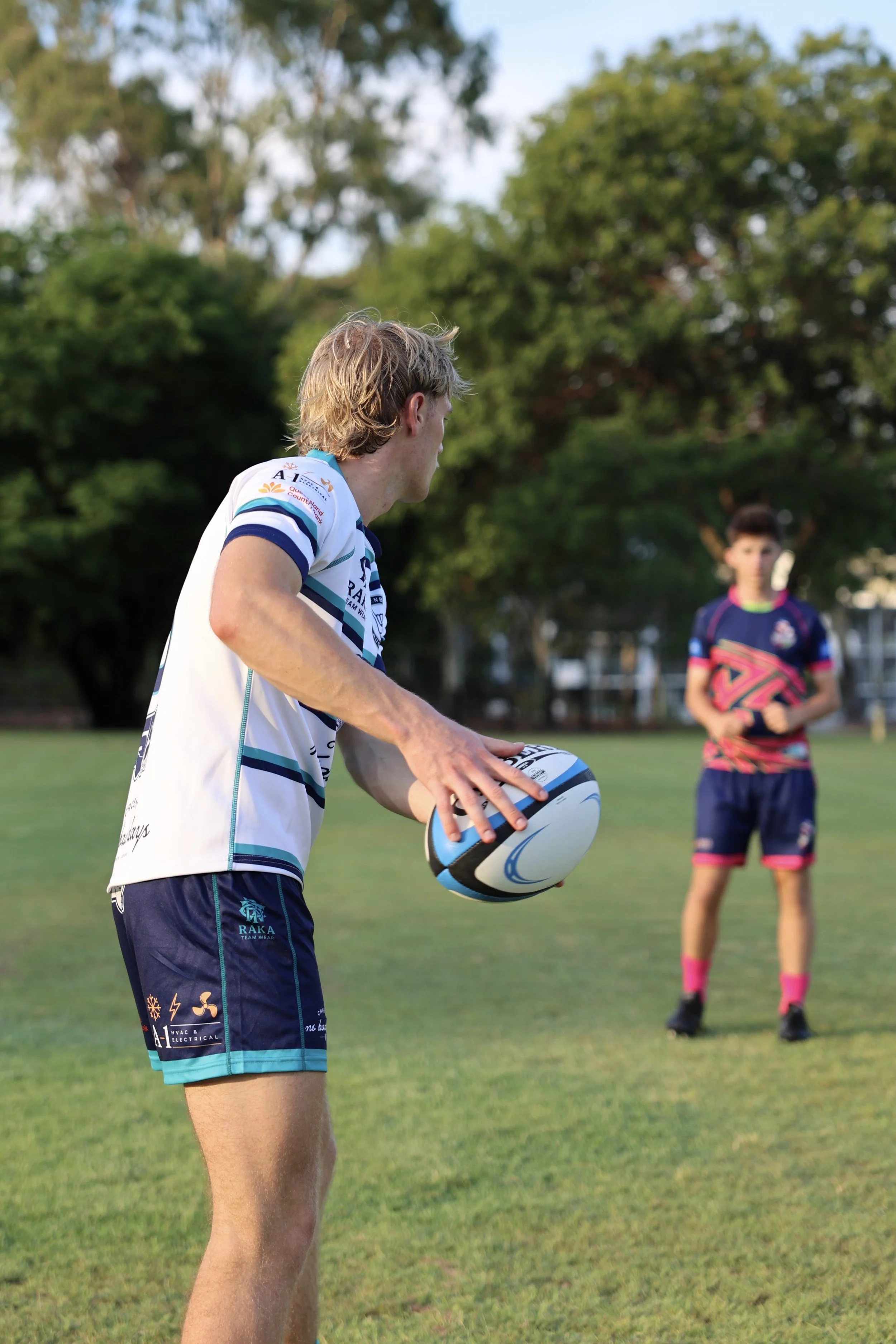 A rugby player in a white and blue jersey prepares to kick a rugby ball on a grassy field with another player in the background.