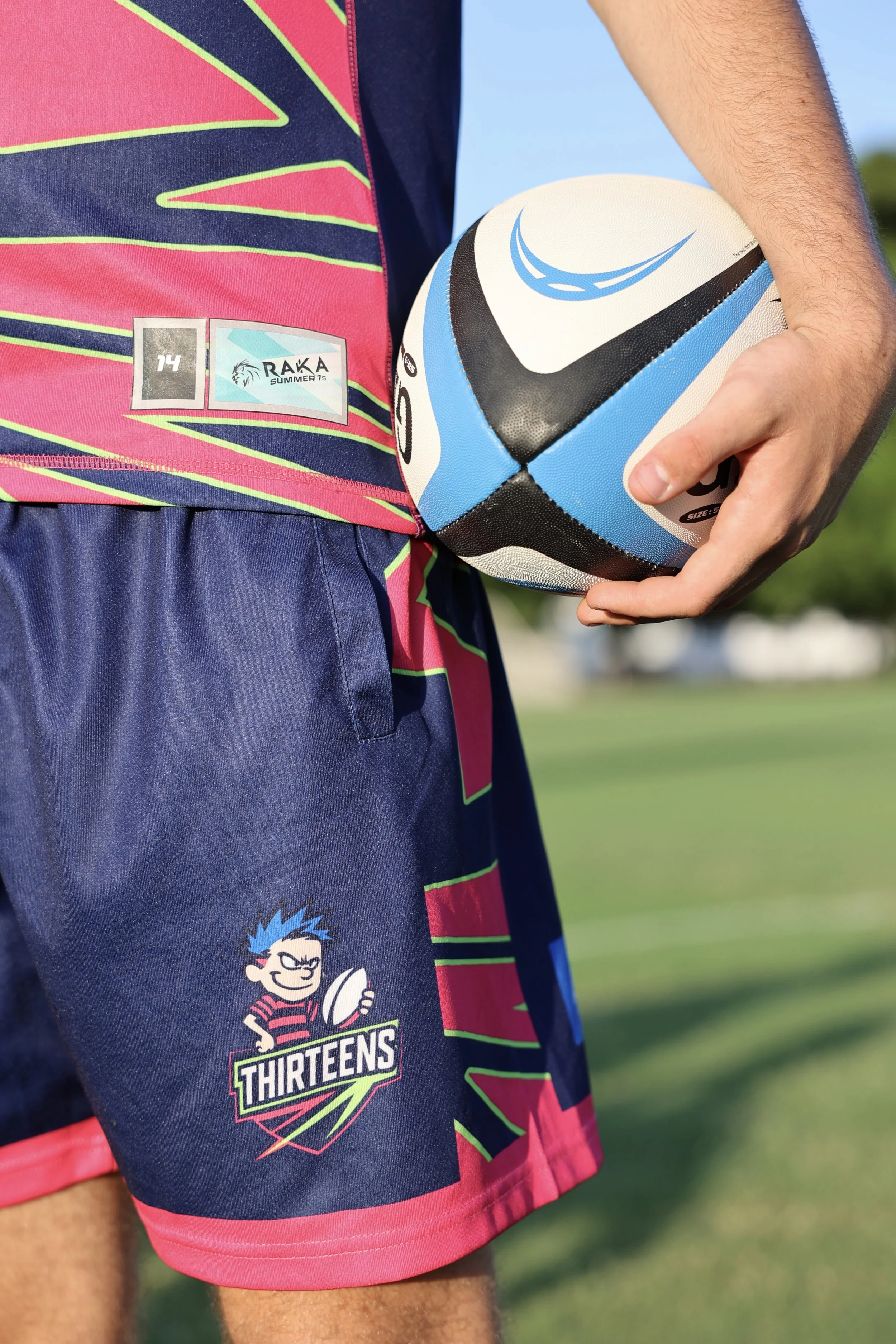 Close-up of a rugby player holding a rugby ball on a field, wearing colorful sports shorts and a jersey with logos, including Thirteens and RAKA Summer T.