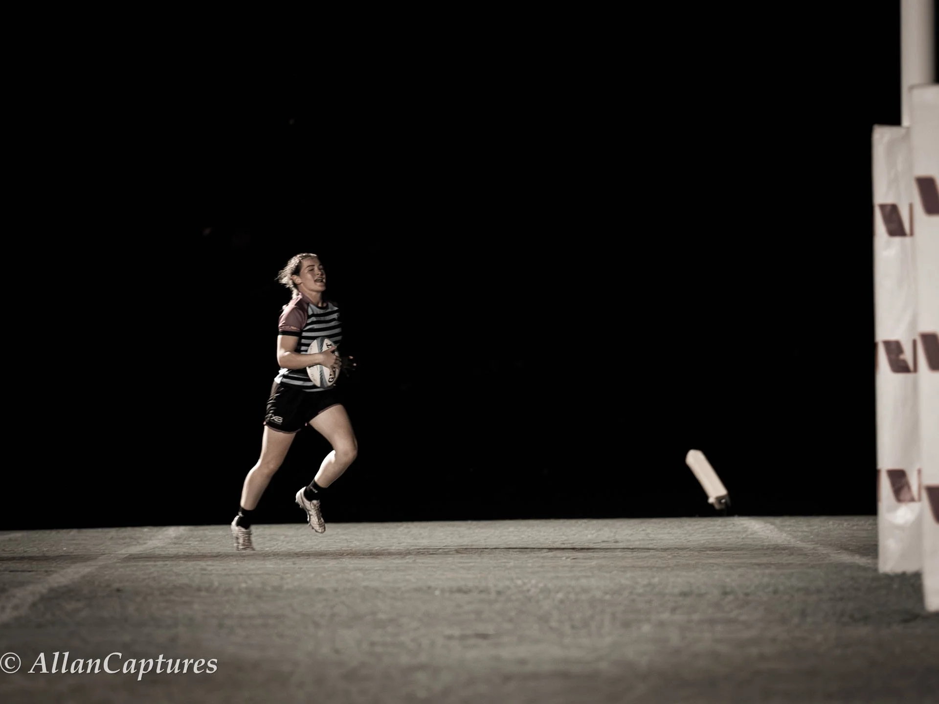 A female rugby player running with a rugby ball on a dark field, with a goal post in the background.