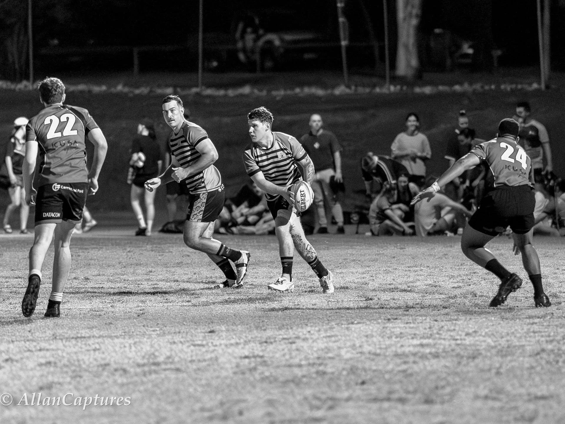 A black and white photo of rugby players on the field, with one player holding the ball, while others are in motion, and spectators in the background.