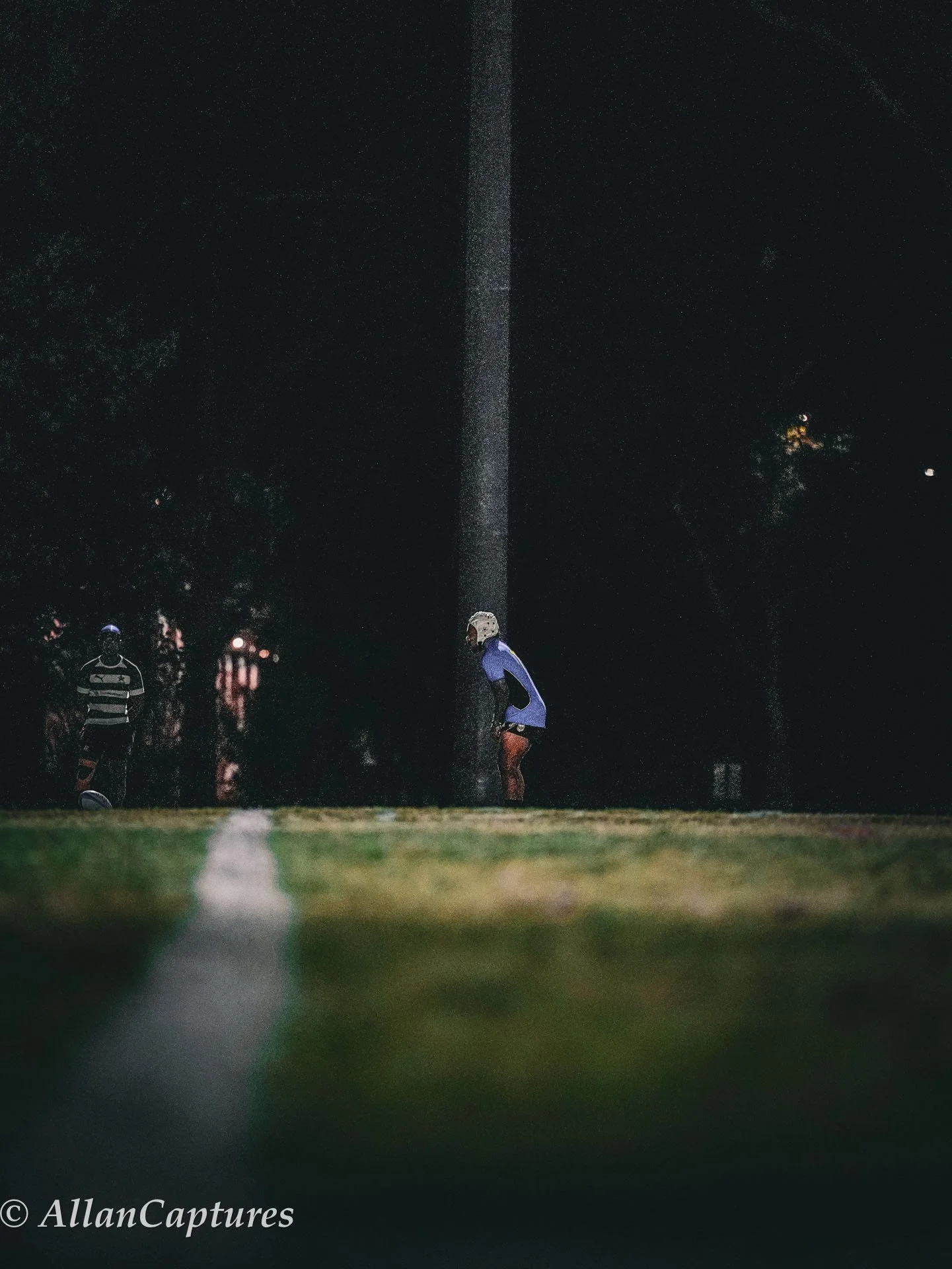A person wearing a helmet and blue jacket stands on a football field at night, with water or spray falling from above, illuminated against a dark background. Another person is visible in the background on the left side of the image.