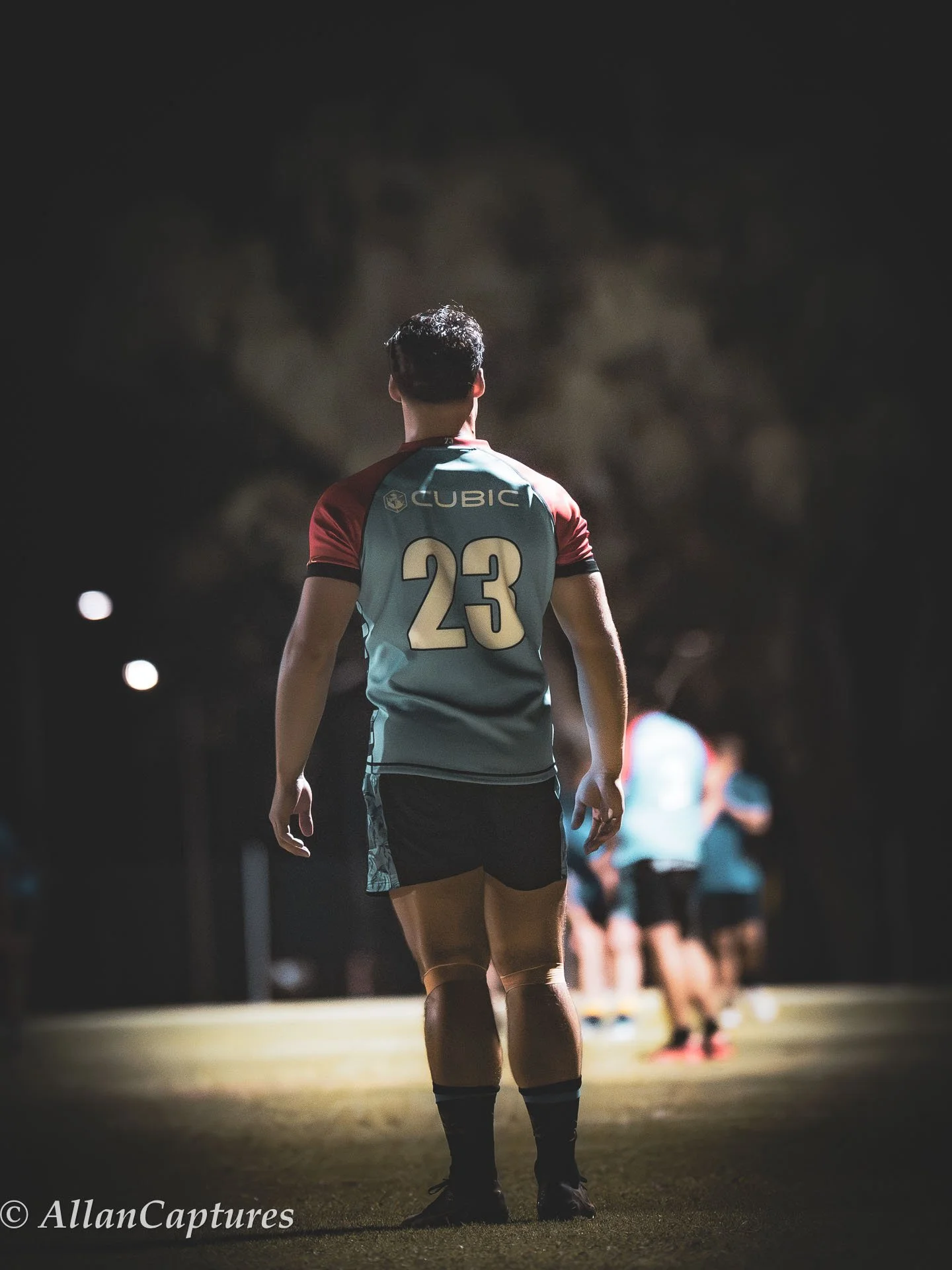 A rugby player with jersey number 23 standing on a field at night, facing away from the camera, with other players in the background.