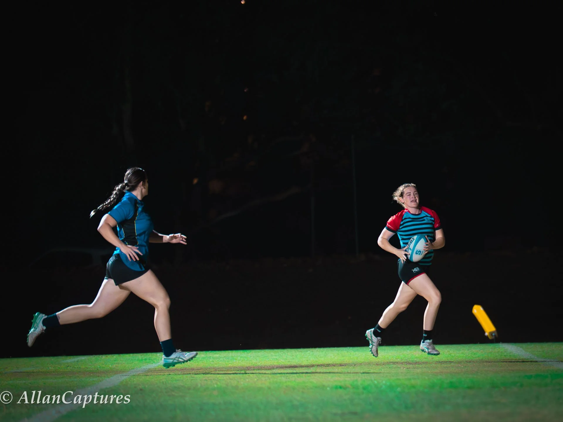 Two female rugby players running on a rugby field at night, one holding a rugby ball, with a dark background.