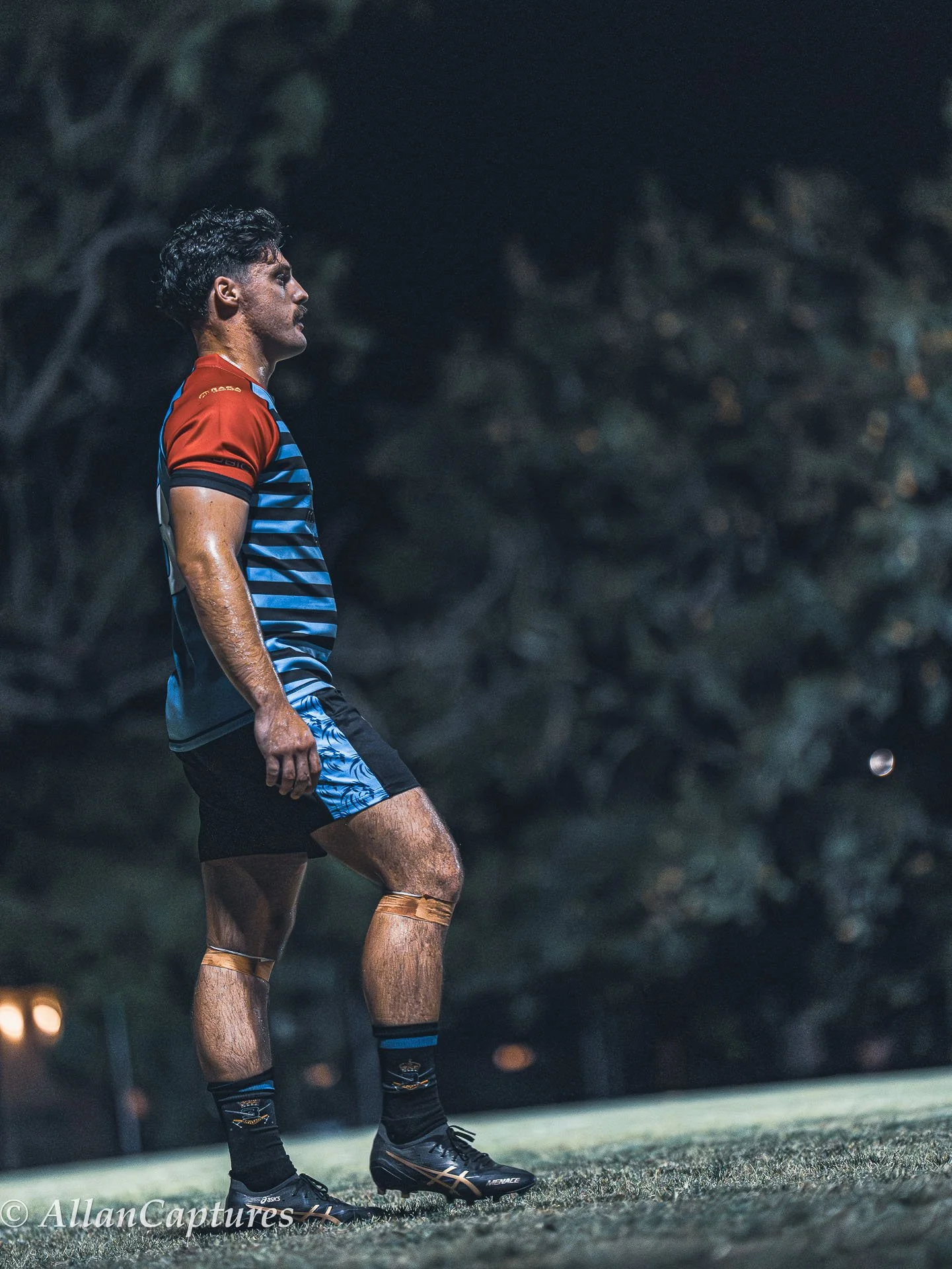 A male rugby player standing on a field at night, wearing a blue and black striped jersey with red sleeves, black shorts, black socks, and rugby boots, with a rugby ball in the distance.