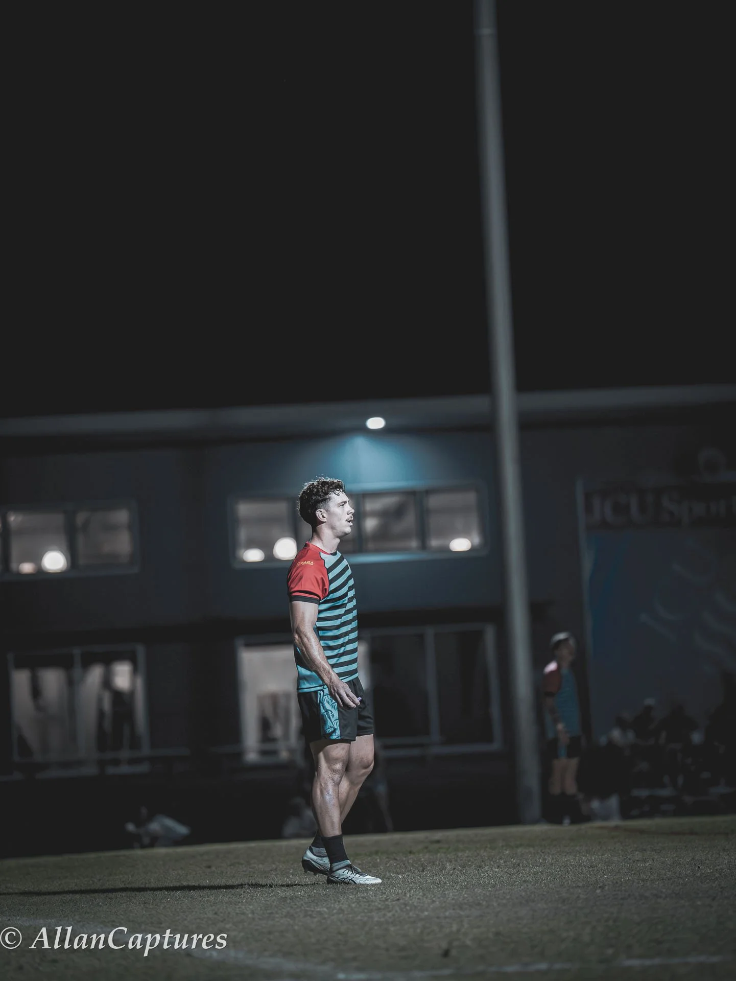 A young male athlete standing on a sports field at night, wearing a striped athletic shirt and shorts, with a serious expression, under stadium lights.