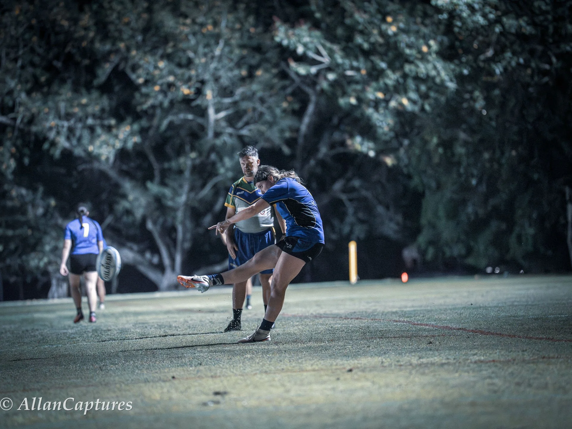 A female rugby player in a blue uniform kicks a rugby ball while a male teammate in a striped jersey watches on a field at night, with other players in the background.