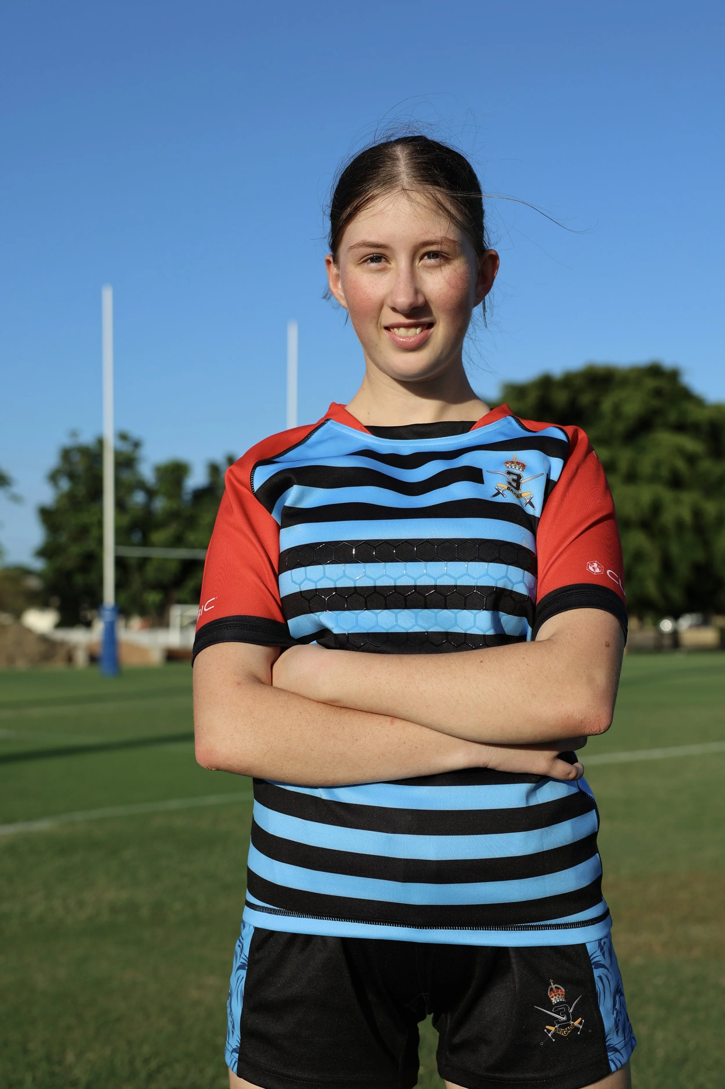 Young female rugby player standing on a field with arms crossed, wearing a striped blue and black jersey with red sleeves, under a clear blue sky.