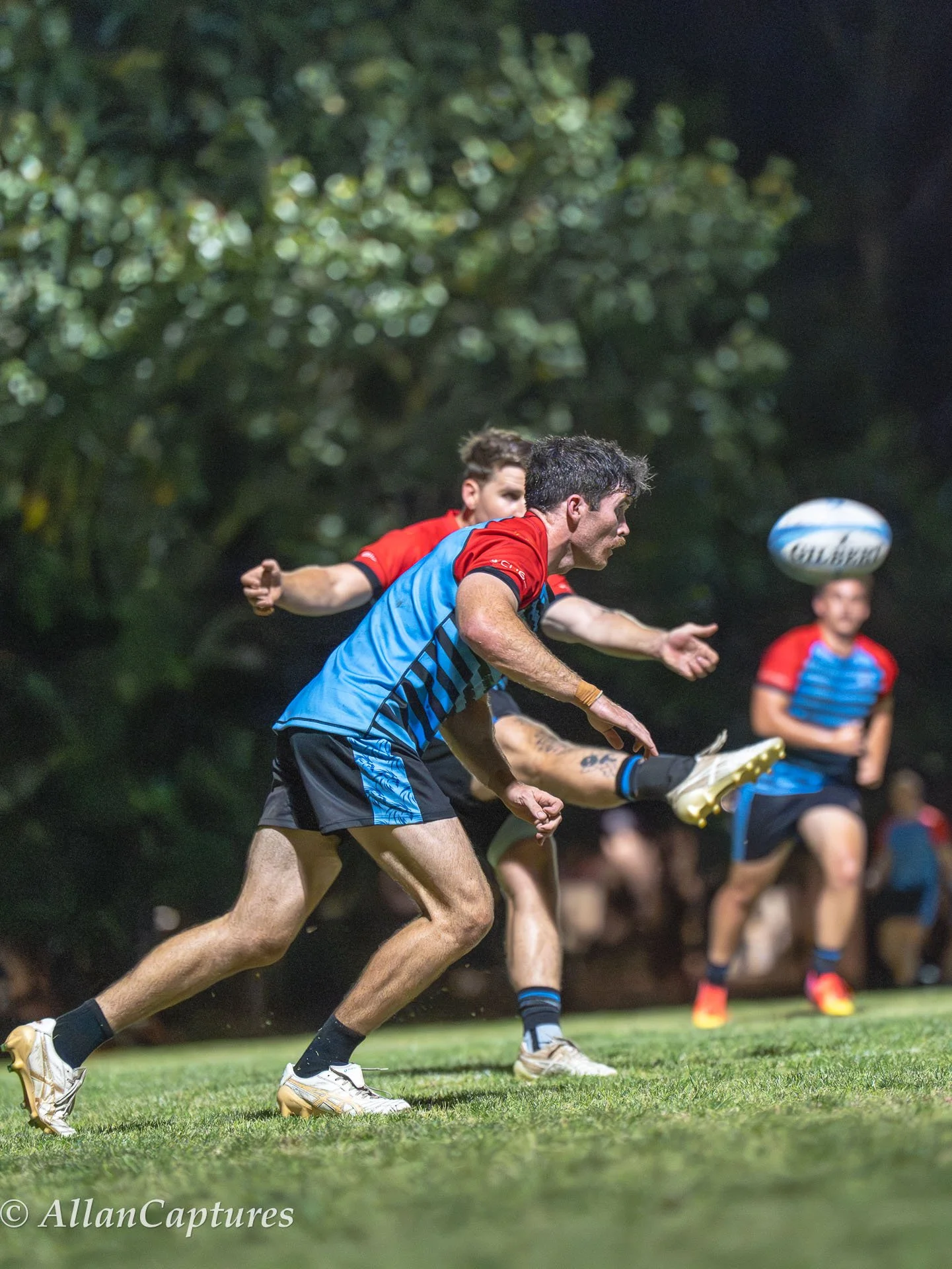 Rugby players in action during a night game, one player passing the ball while others run on a grass field with trees in the background.