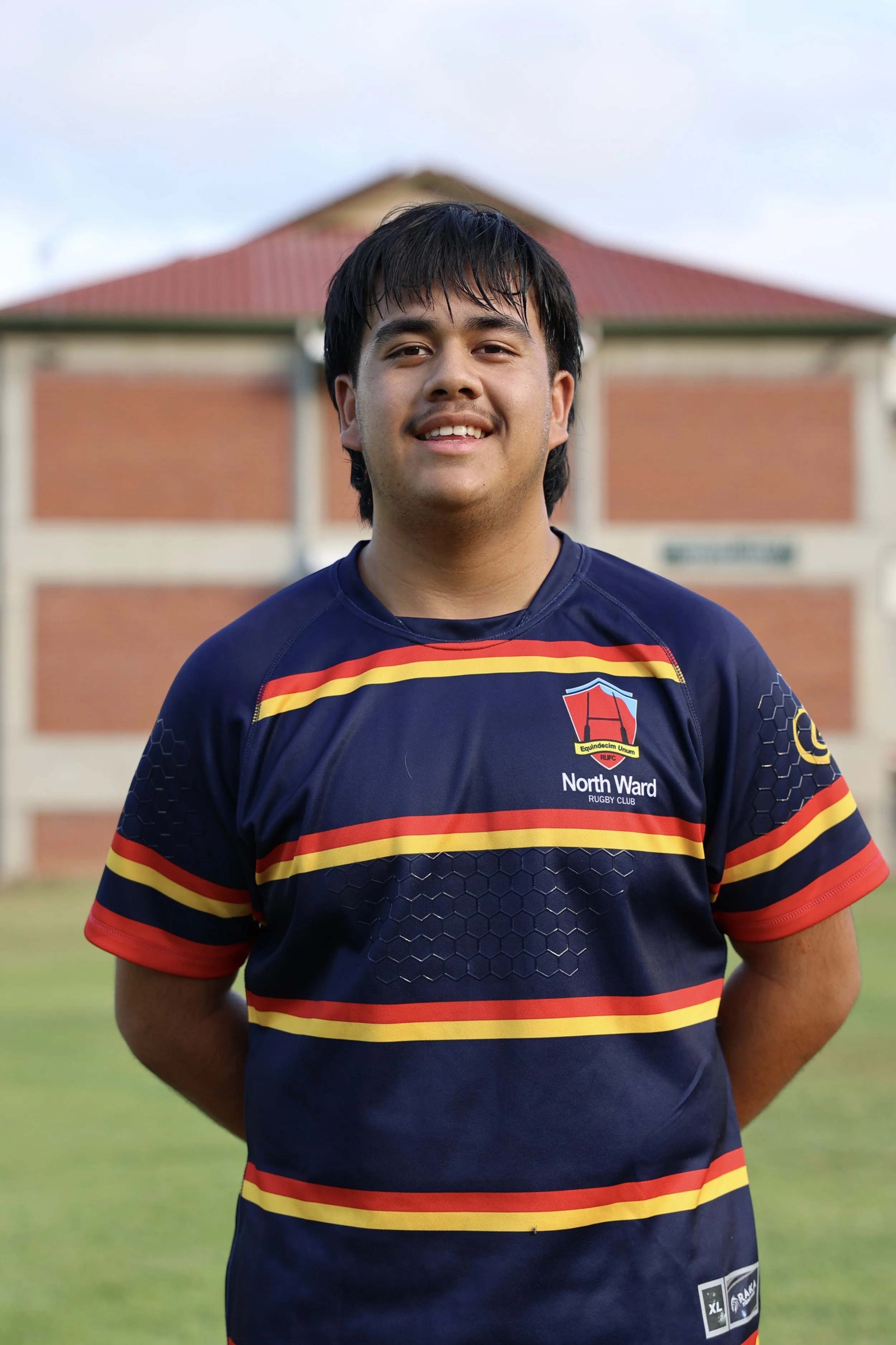 Young man in a blue rugby jersey standing outdoors on grass, smiling, with a brick building in the background.