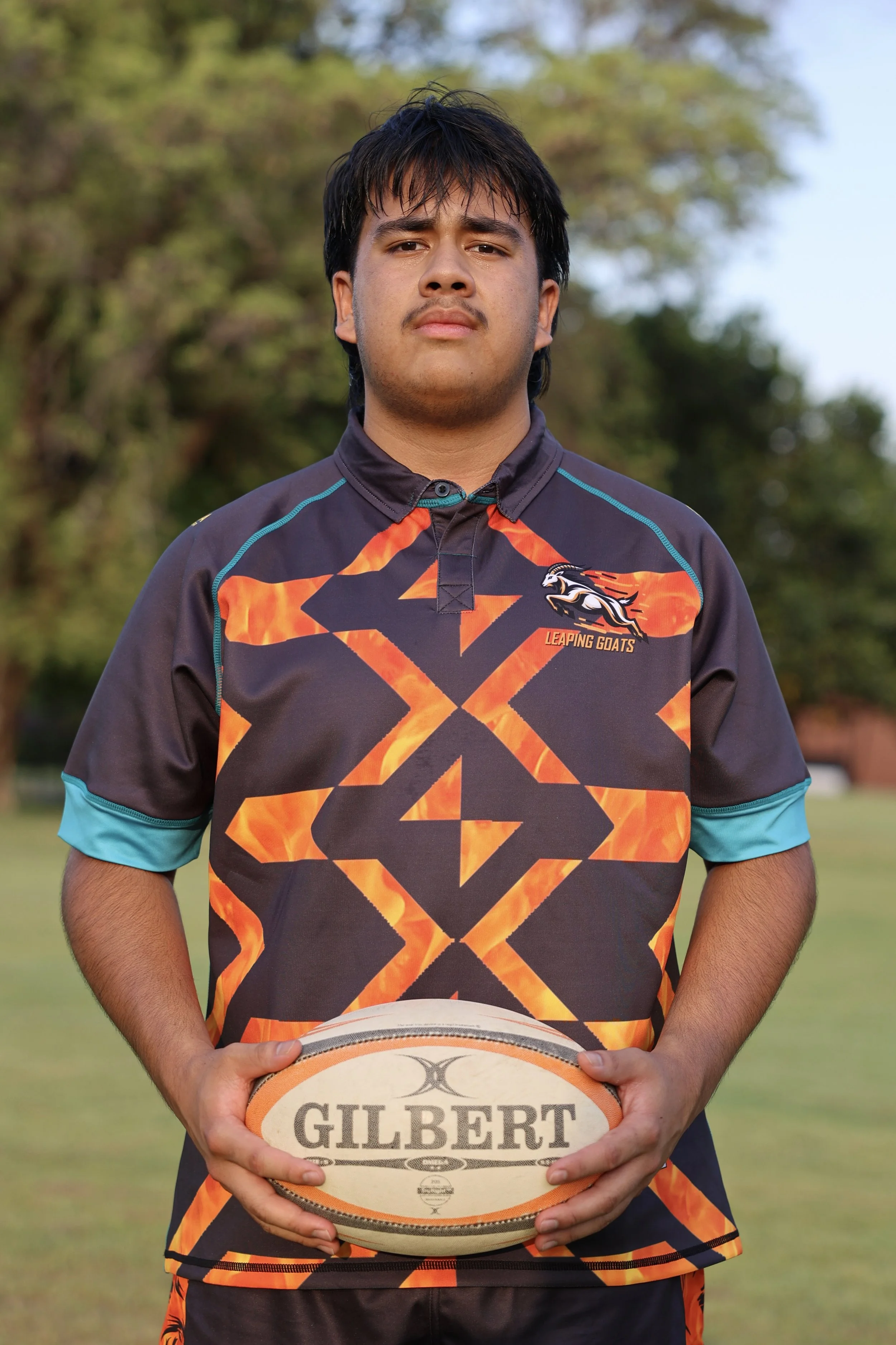 Young rugby player holding a Gilbert rugby ball outdoors, wearing a black and orange sports jersey with blue accents and a logo of a leaping goat.