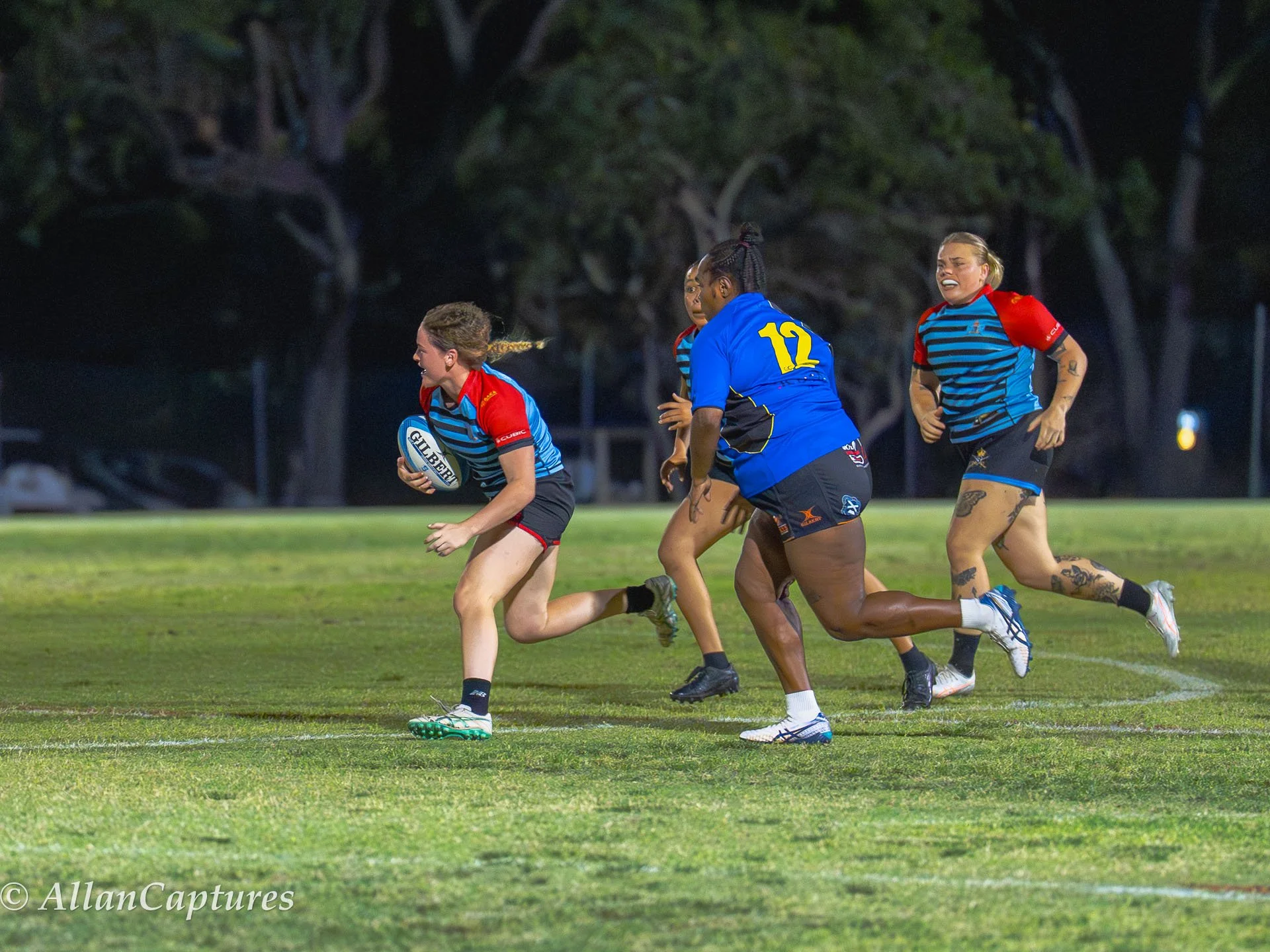 Women playing rugby on a field at night, with one player running with the ball while others chase behind.