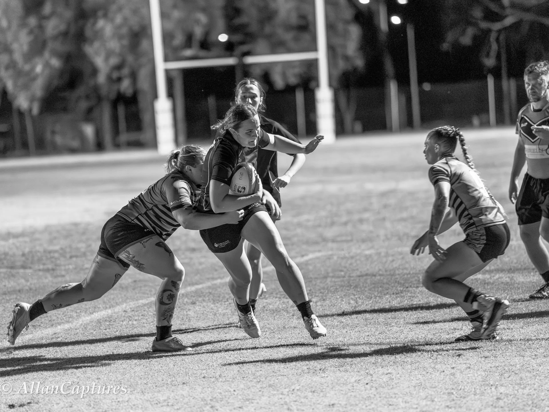 Black and white photo of women playing rugby, with one woman carrying the ball while others attempt to tackle or block her on a grassy field at night.