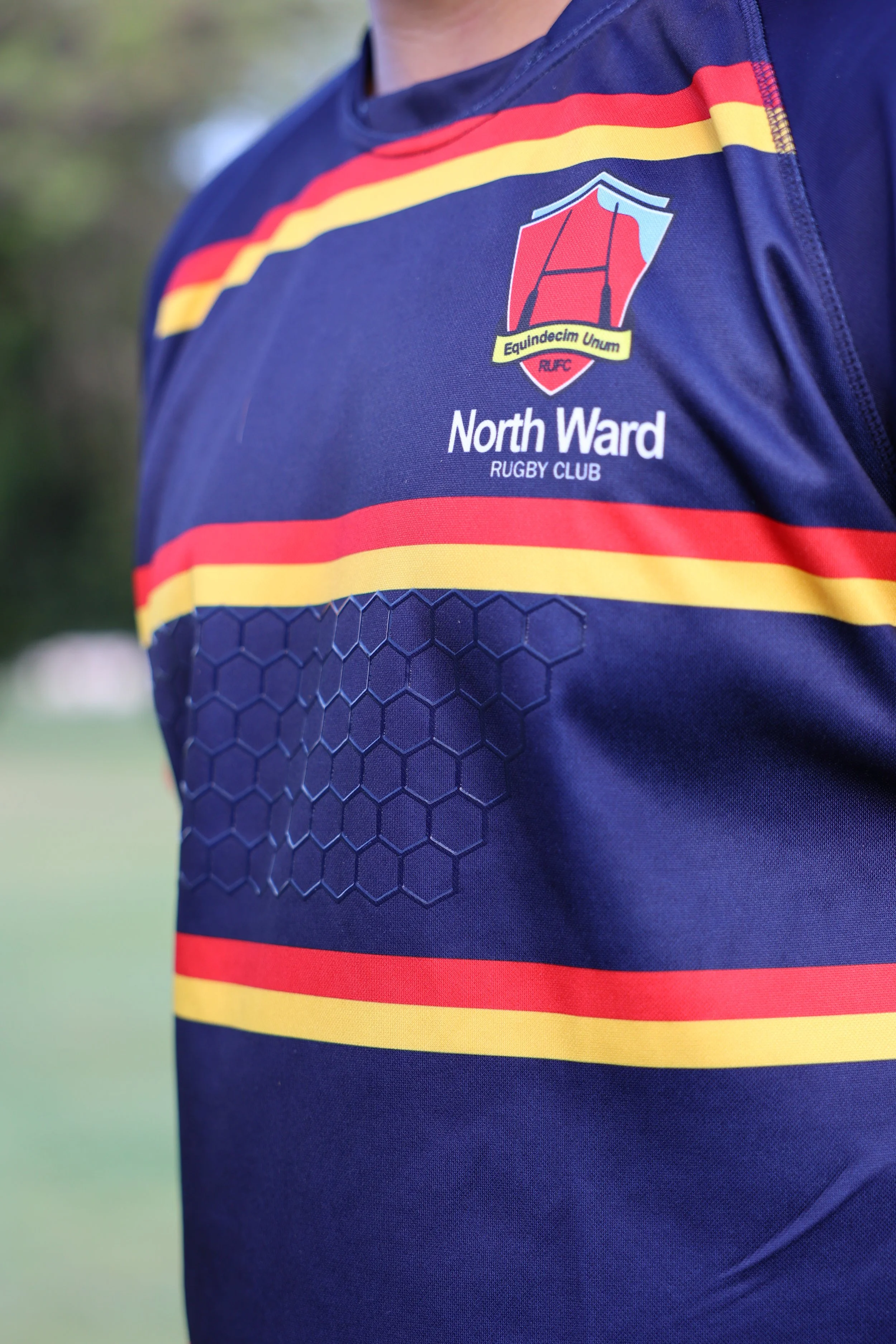 Close-up of a rugby jersey with the North Ward Rugby Club logo and text, featuring red and yellow stripes on a blue background.