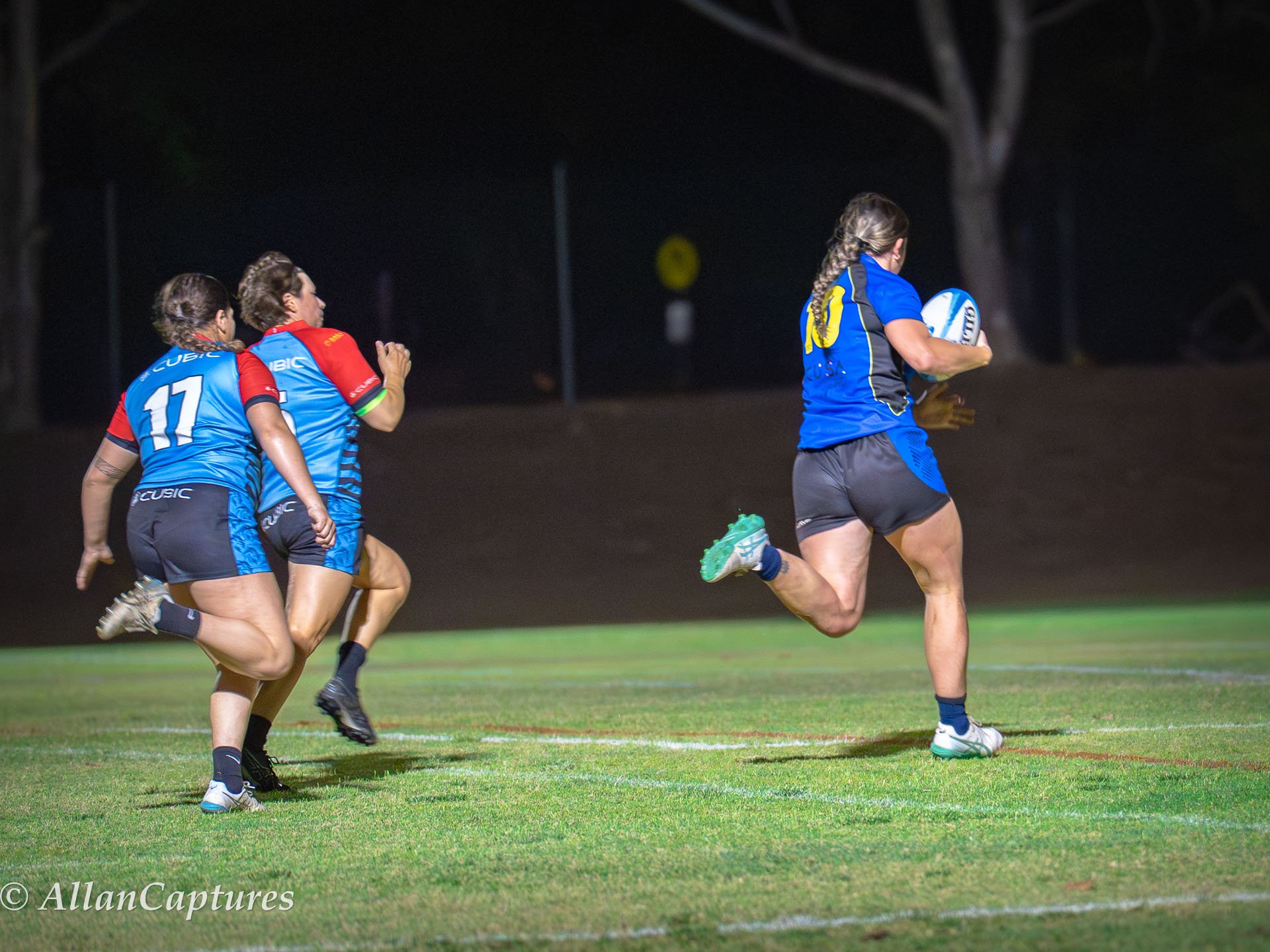 Four women playing rugby on a field at night, with three women chasing a woman running with the ball.