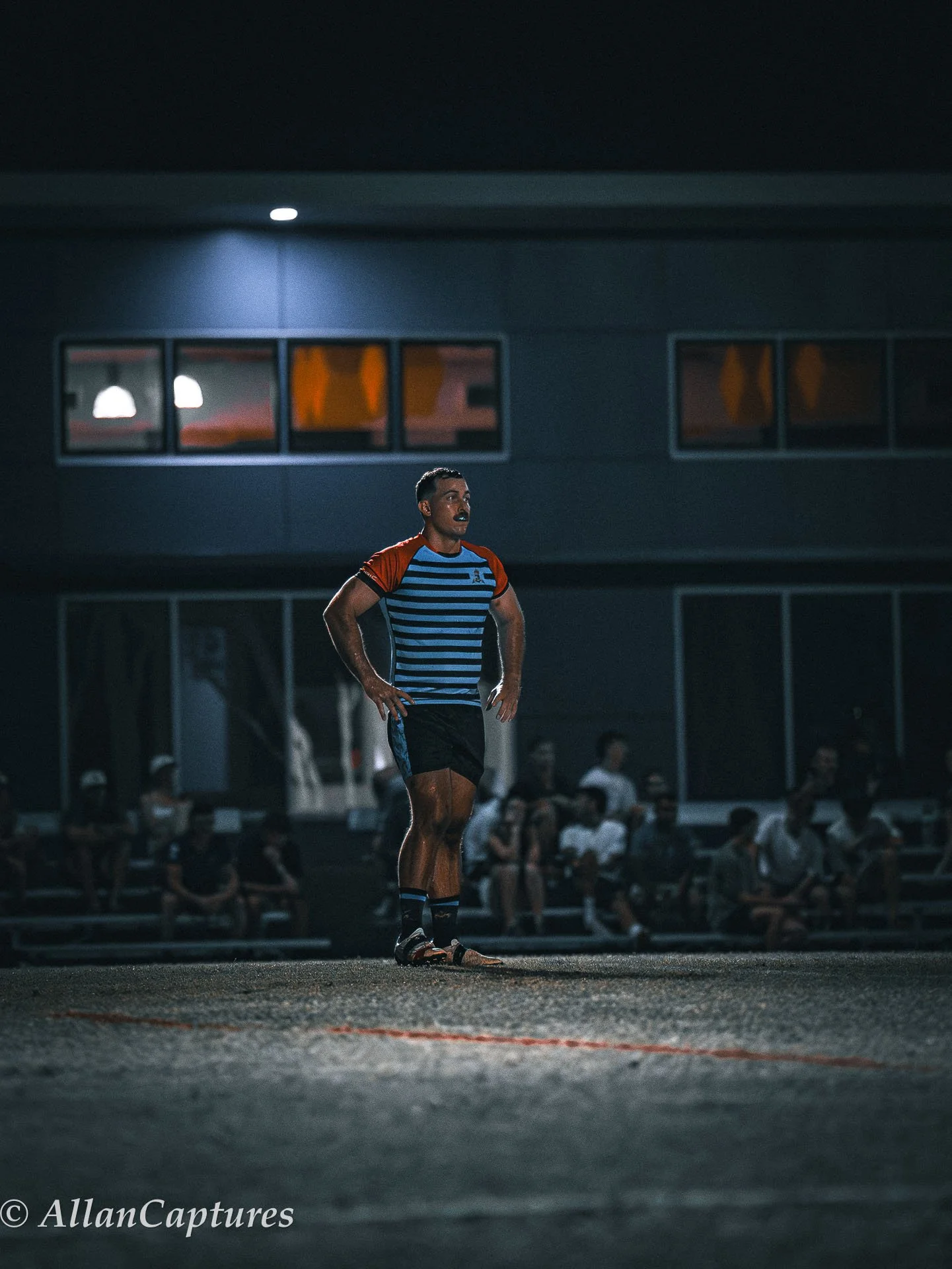A rugby player stands on the field under bright stadium lights during a night game, with spectators in the background.