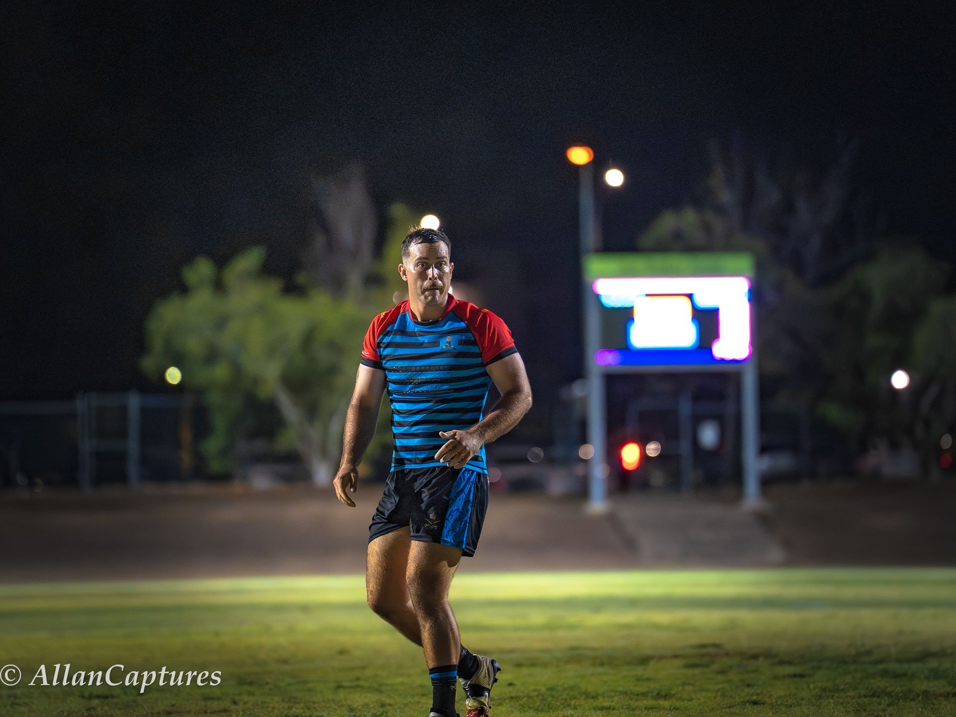 A man jogging on a sports field at night, wearing a blue and red athletic shirt and shorts, with a scoreboard and trees in the background.