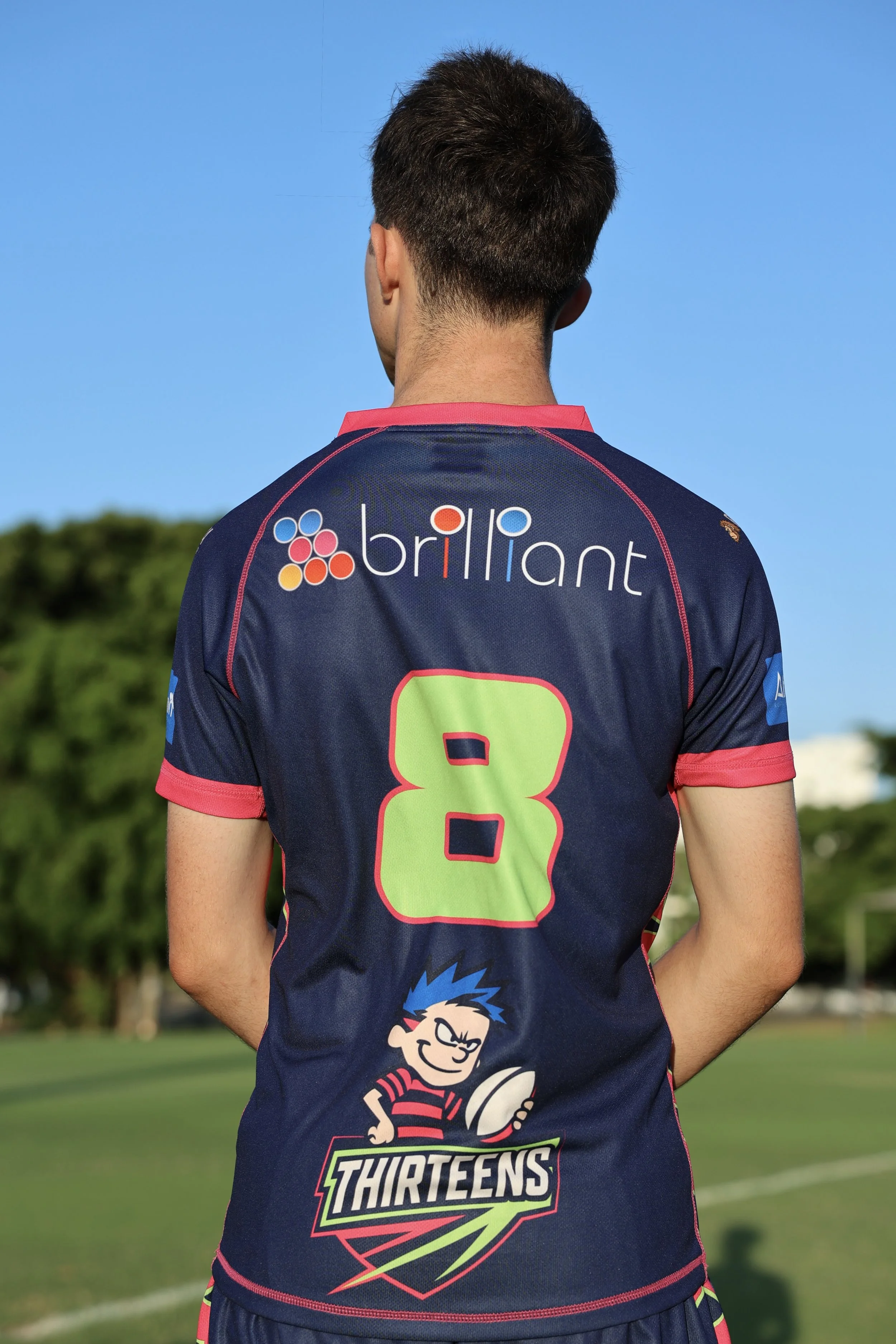 Back of a young male rugby player wearing a navy blue jersey with pink accents, number 8 in bright green, standing outdoors on a rugby field with trees and blue sky in the background.