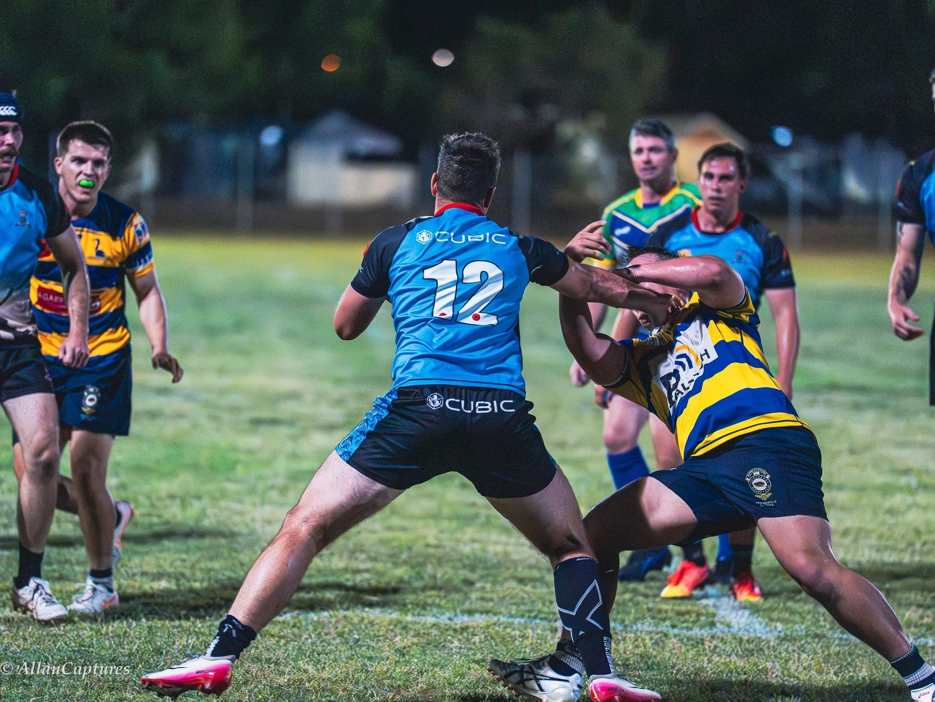 Rugby players engaged in a tackle during a match at night on a grass field, with others watching and some in the background.