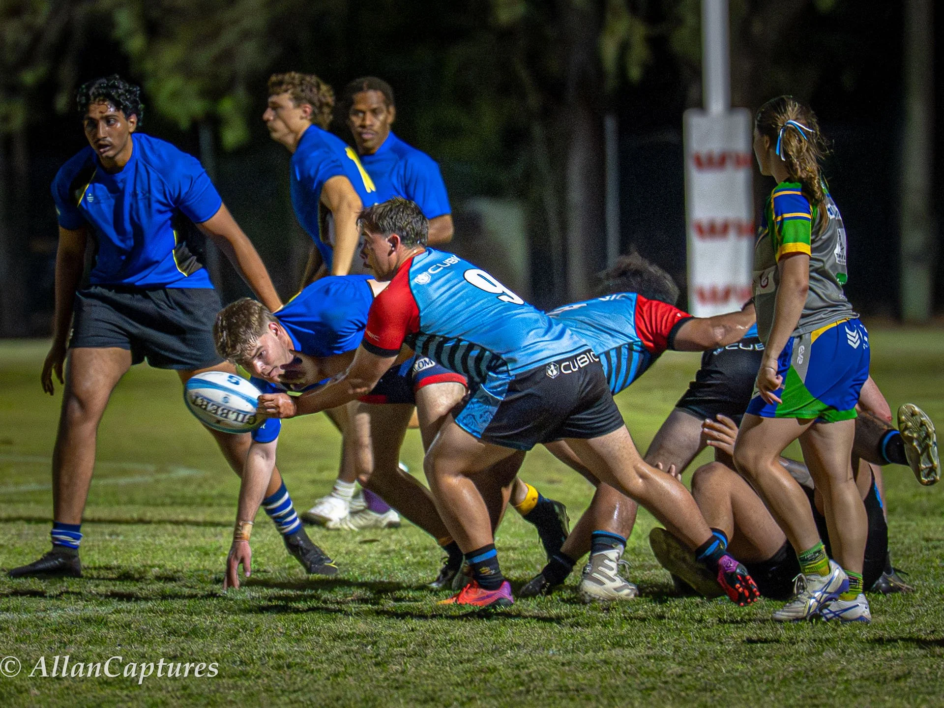 Rugby players in blue and black jerseys competing for the ball during a match at night on a grass field.
