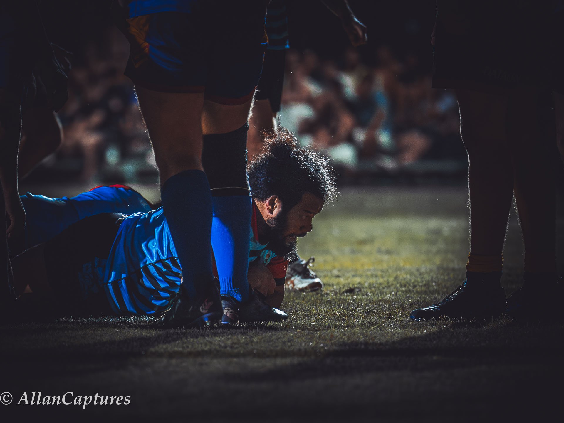 A rugby player wearing a blue uniform is lying on the ground, being tackled or helped by others during a match. The scene is dimly lit, with an audience blurred in the background.