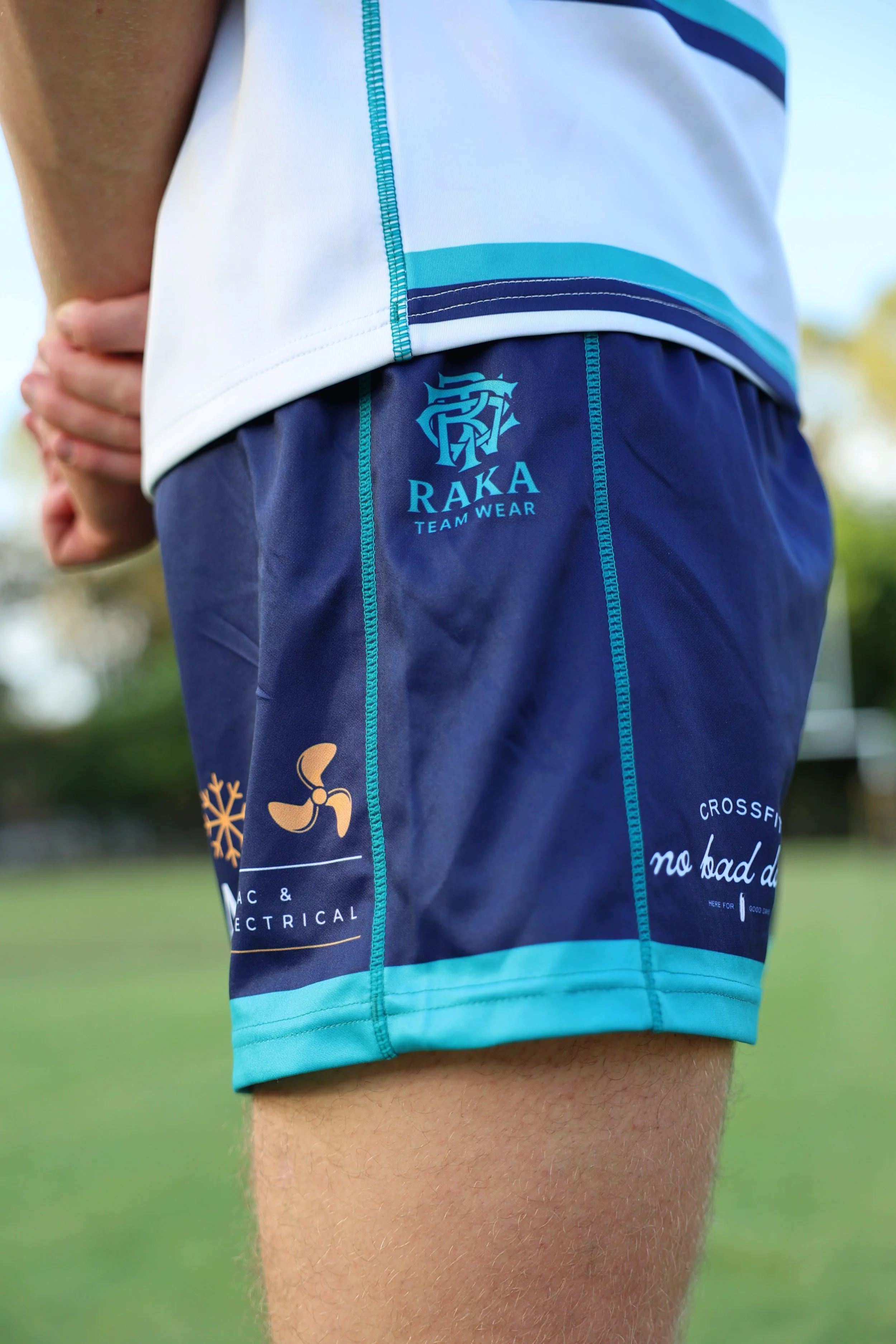 Close-up of a person wearing navy blue athletic shorts with the RAKA Team Wear logo and a graphic of a propeller on the side. Part of a white sports jersey with blue and turquoise stripes is visible. The person is outdoors, standing on a grassy field