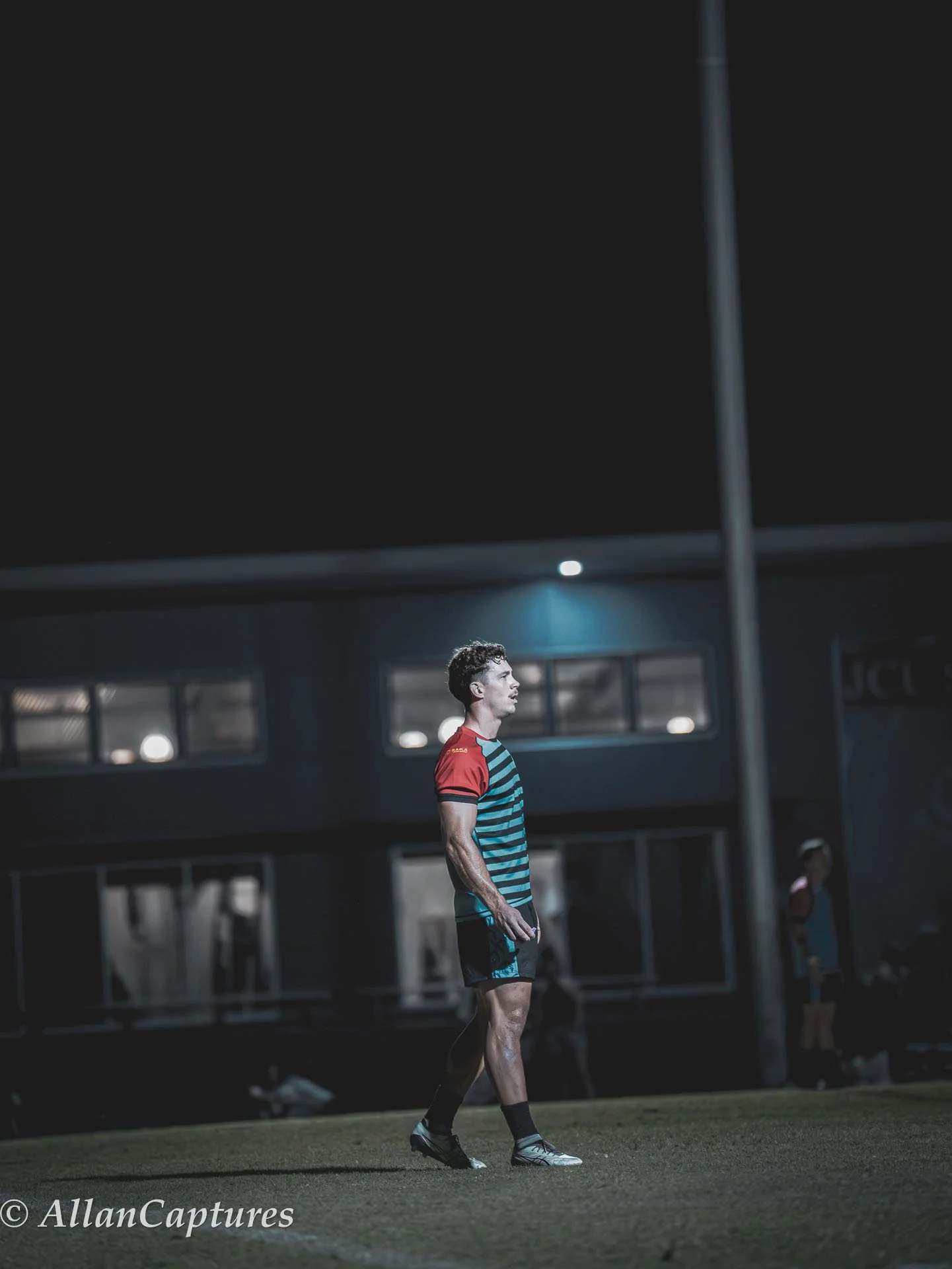 A male soccer player standing on a football field at night, illuminated by stadium lights, wearing a blue and black striped jersey with red sleeves, black shorts, and cleats.