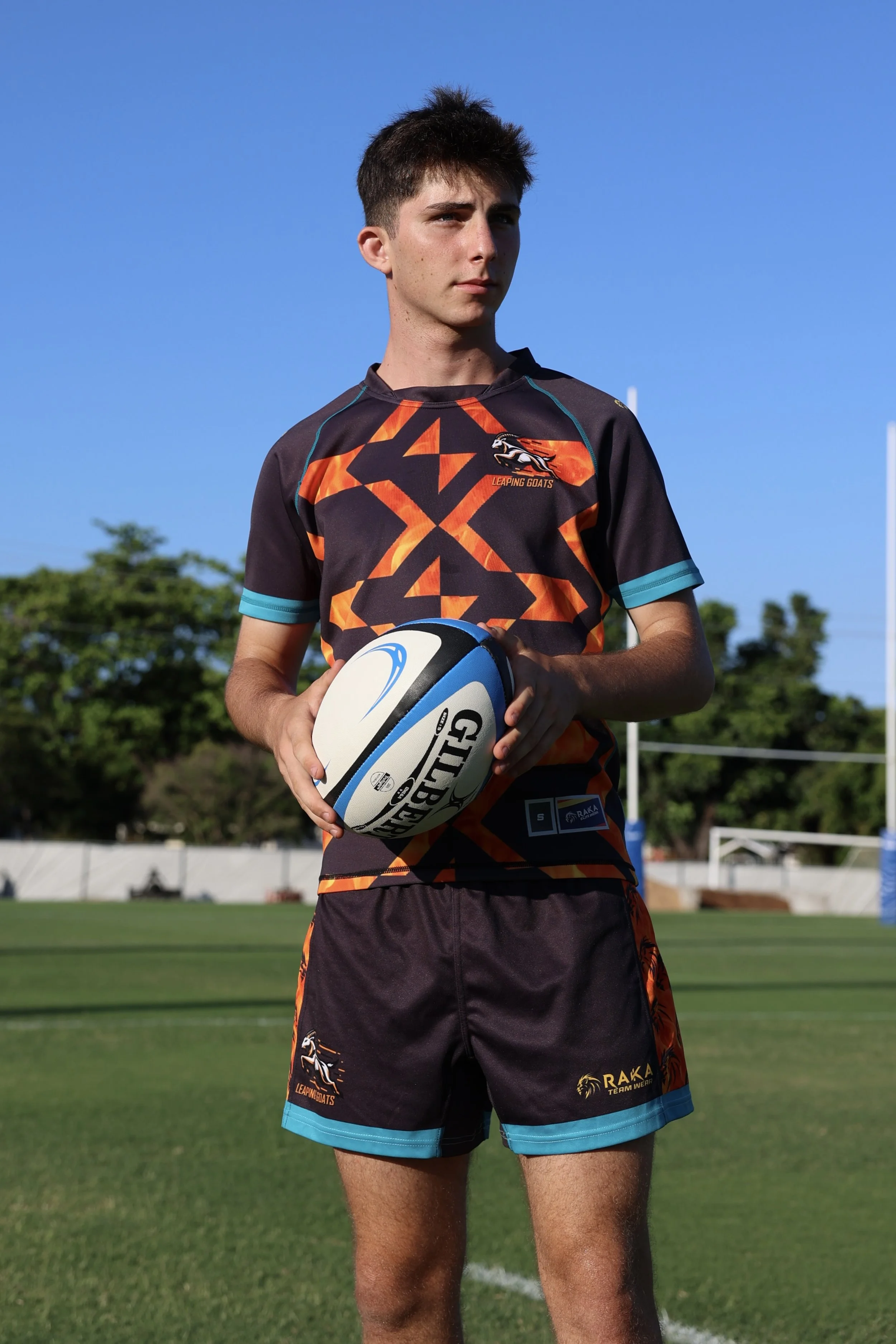 A young male rugby player holding a rugby ball on a rugby field, wearing a black and orange uniform with blue accents, with trees and blue sky in the background.