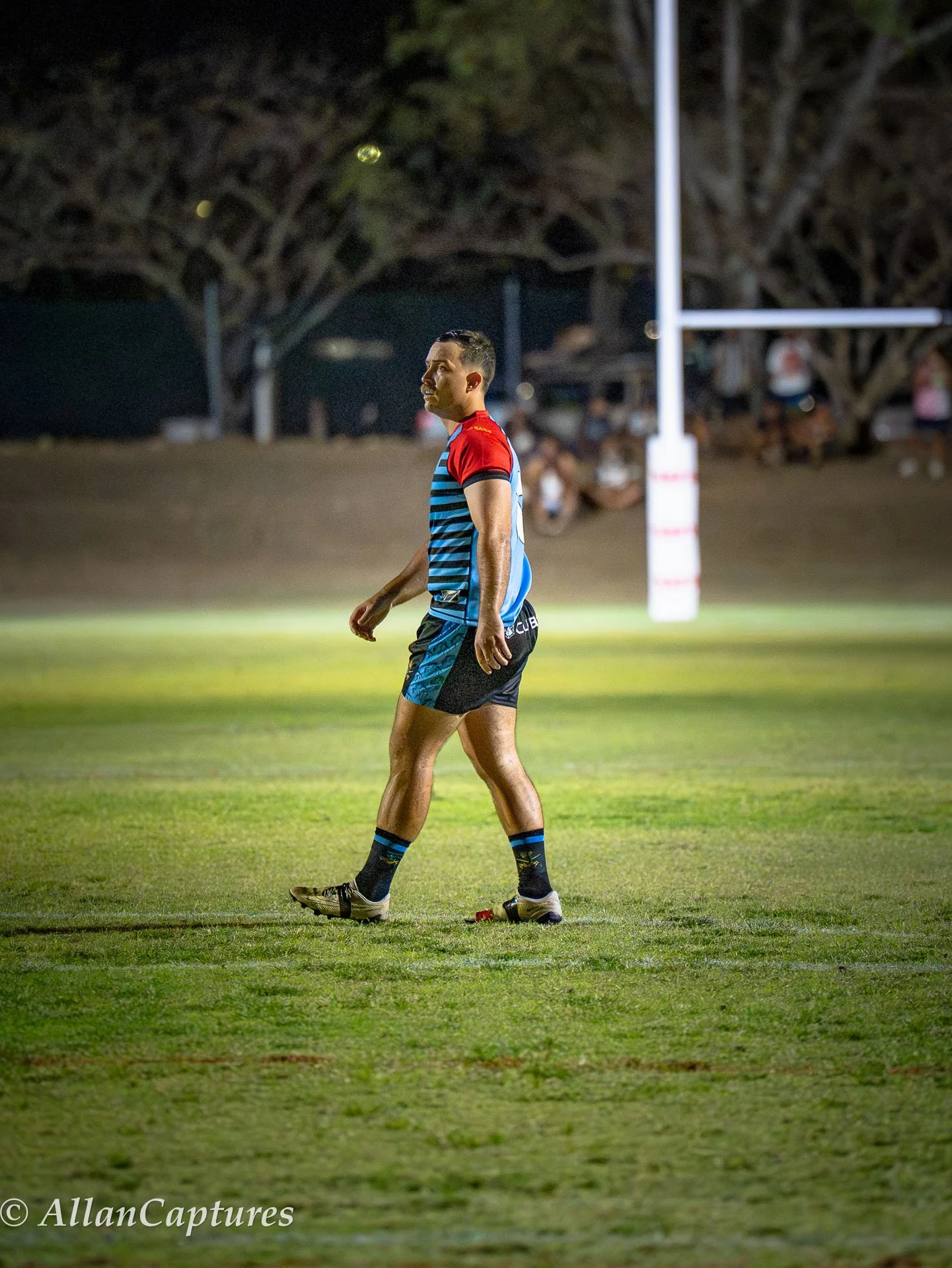 Rugby player standing on the field during a nighttime game, wearing a rugby uniform with red, blue, and black colors, with goalposts in the background.