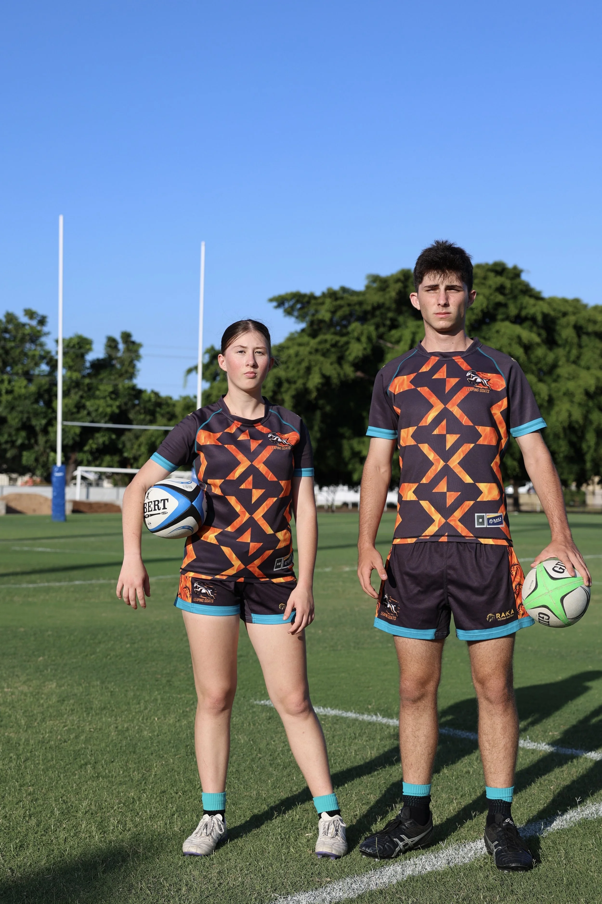 Two rugby players, a woman and a man, standing on a rugby field holding rugby balls, wearing matching team uniforms with orange and black geometric patterns, with goal posts and trees in the background on a sunny day.