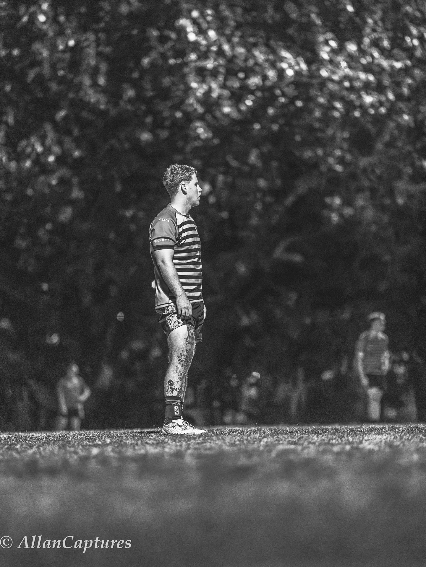 A rugby player stands on the field during a nighttime match, wearing a striped jersey and shorts, with tattoos visible on his legs, and other players in the background.