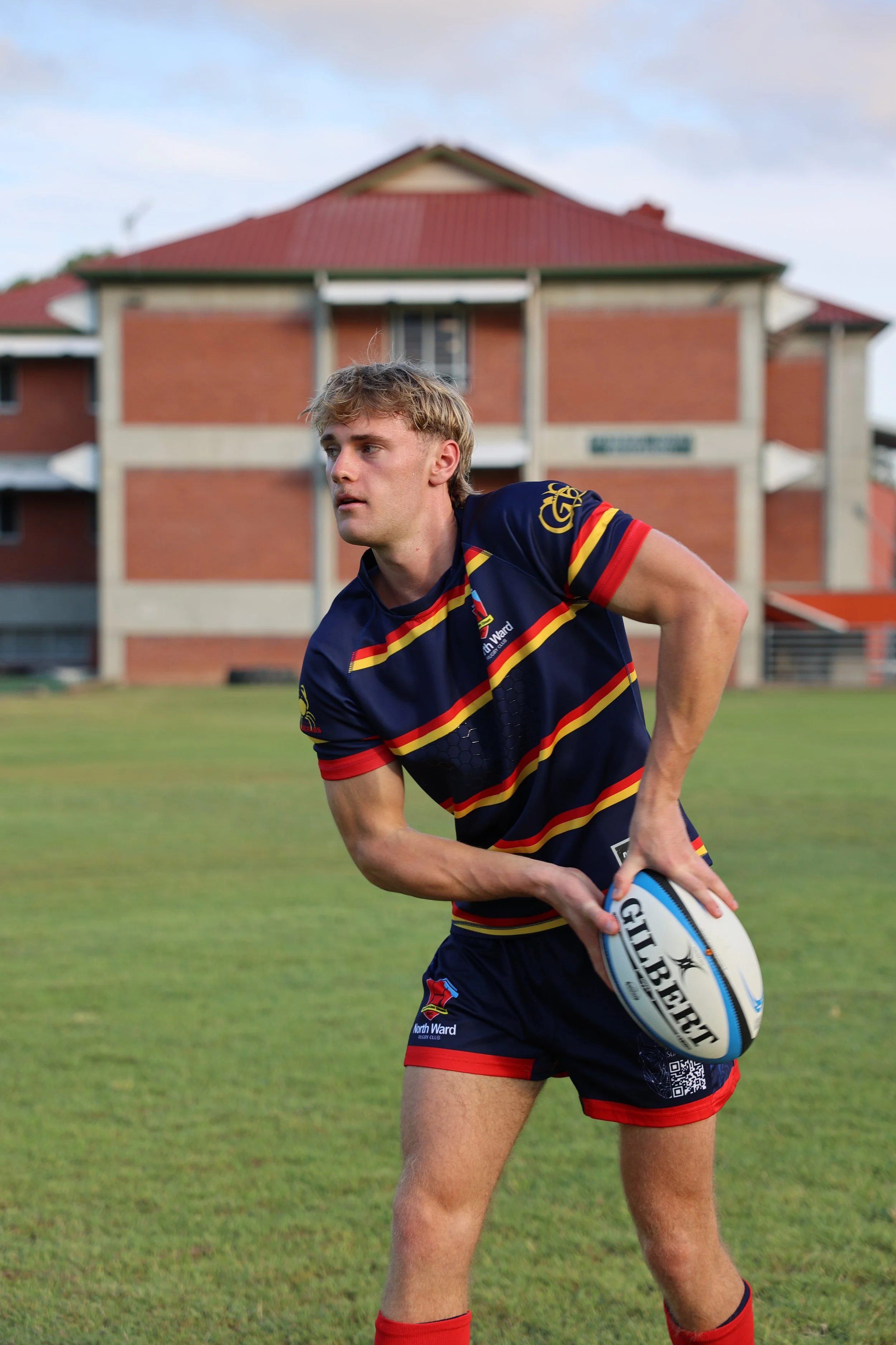 A young woman holding a rugby ball on a grass field with a brick building in the background.