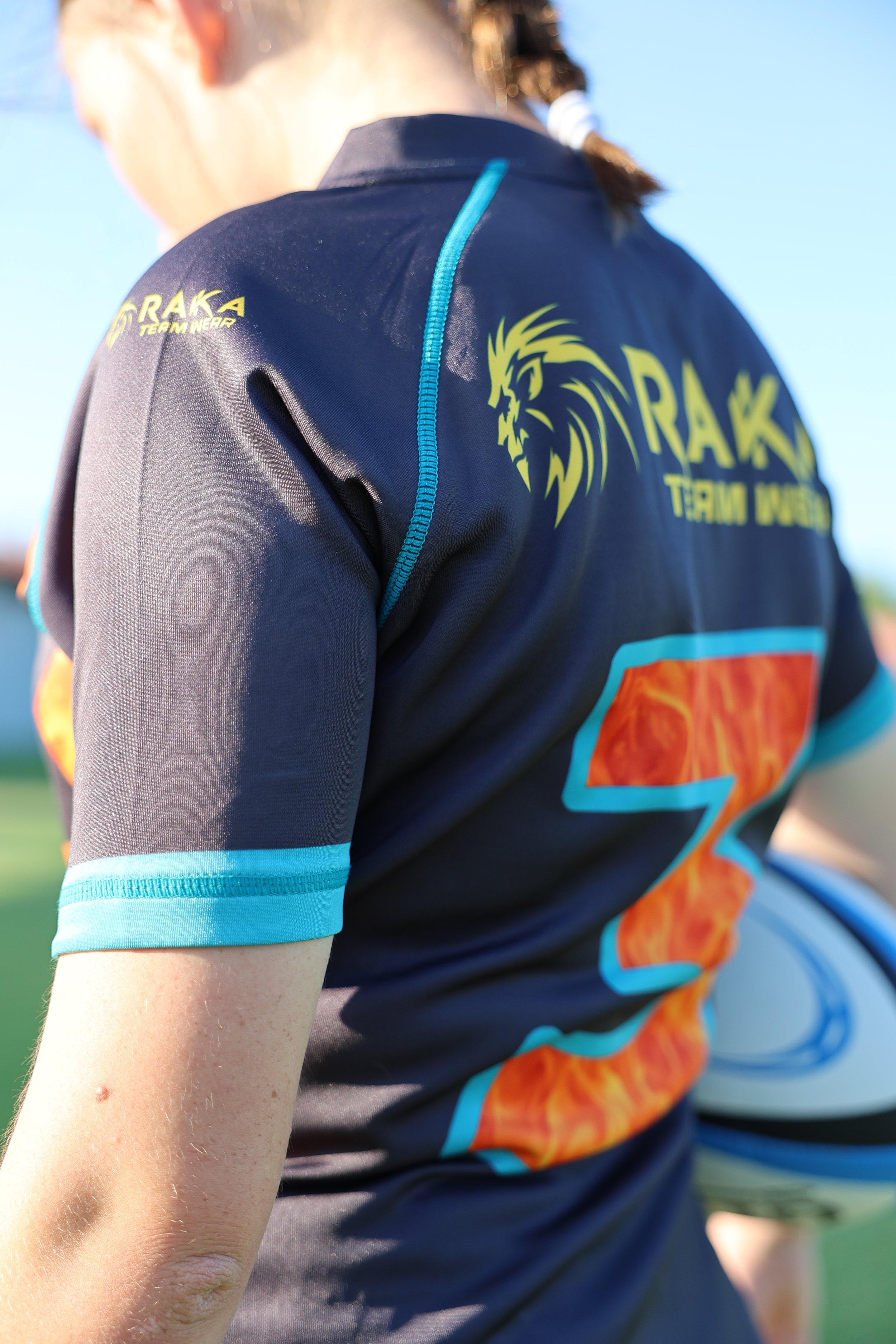 Close-up of a person wearing a black and blue sports jersey with a lion logo and the text 'RAK TEAM WEAR' and a large number '3' on the back, holding a rugby ball on a sunny day.