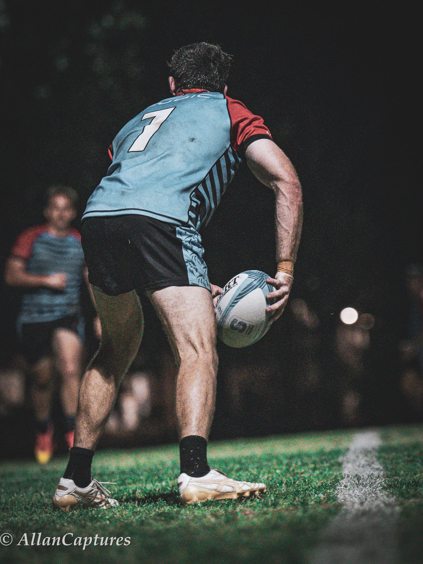 A rugby player in a blue and red jersey about to throw a rugby ball during a night game on a grass field.