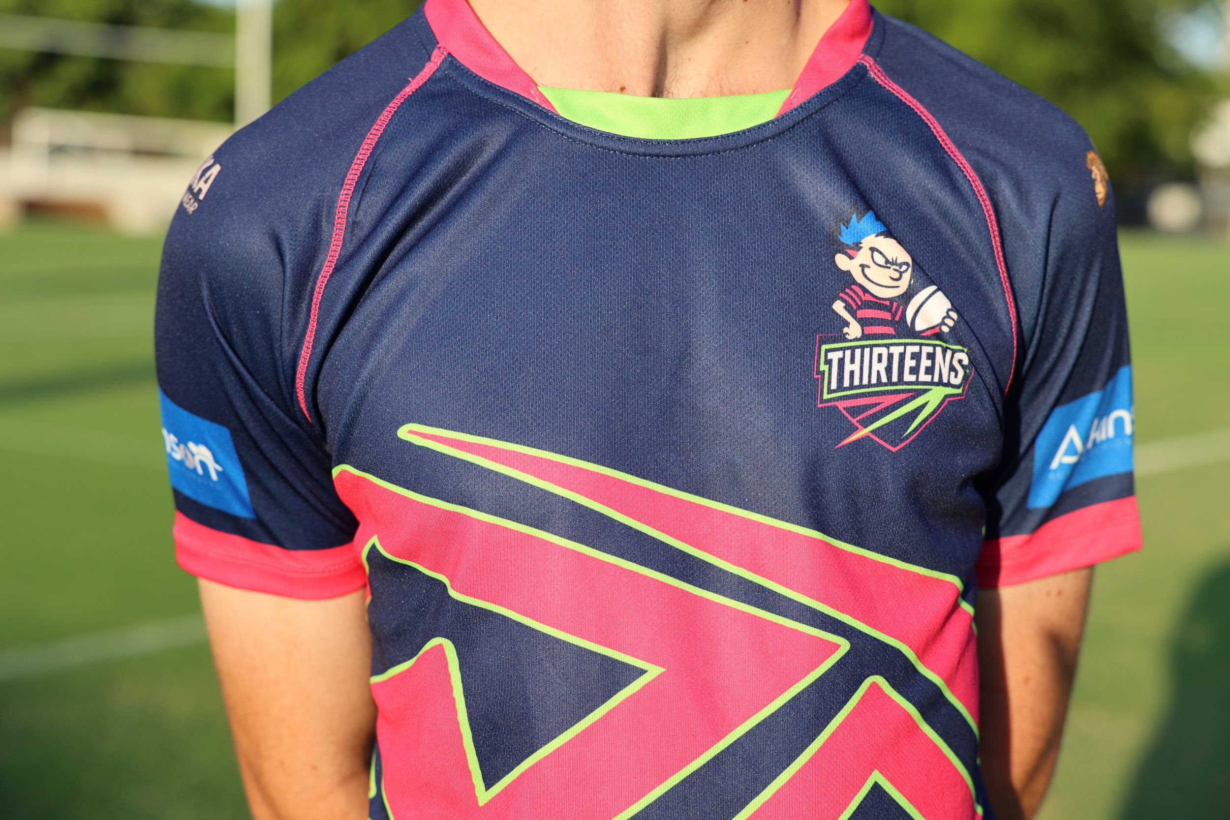 Close-up of a person wearing a navy, pink, and neon yellow rugby jersey on a grassy field. The jersey features a cartoon character logo with the word "THIRTEENS" and aiding sponsor logos on the sleeves.