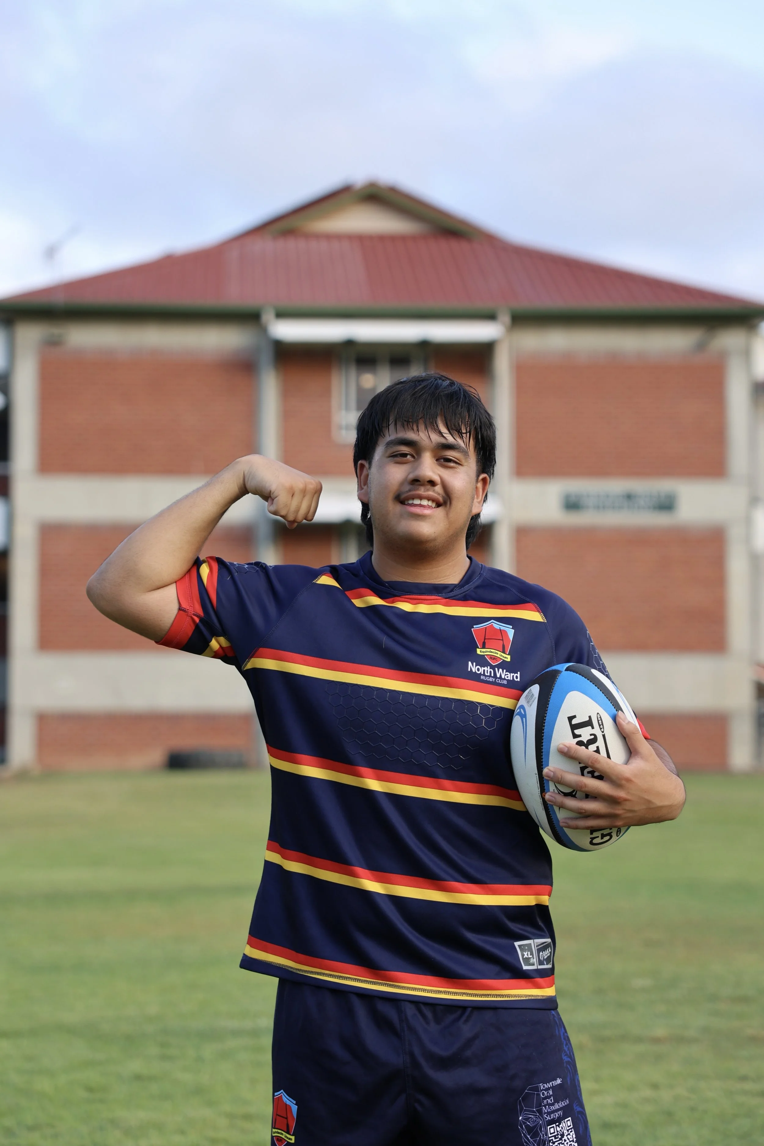 Young man standing on a rugby field holding a rugby ball, flexing his right arm, and smiling. He is wearing a blue sports jersey with red and yellow stripes and the logo of North Ward Rugby Club. A brick building is in the background.