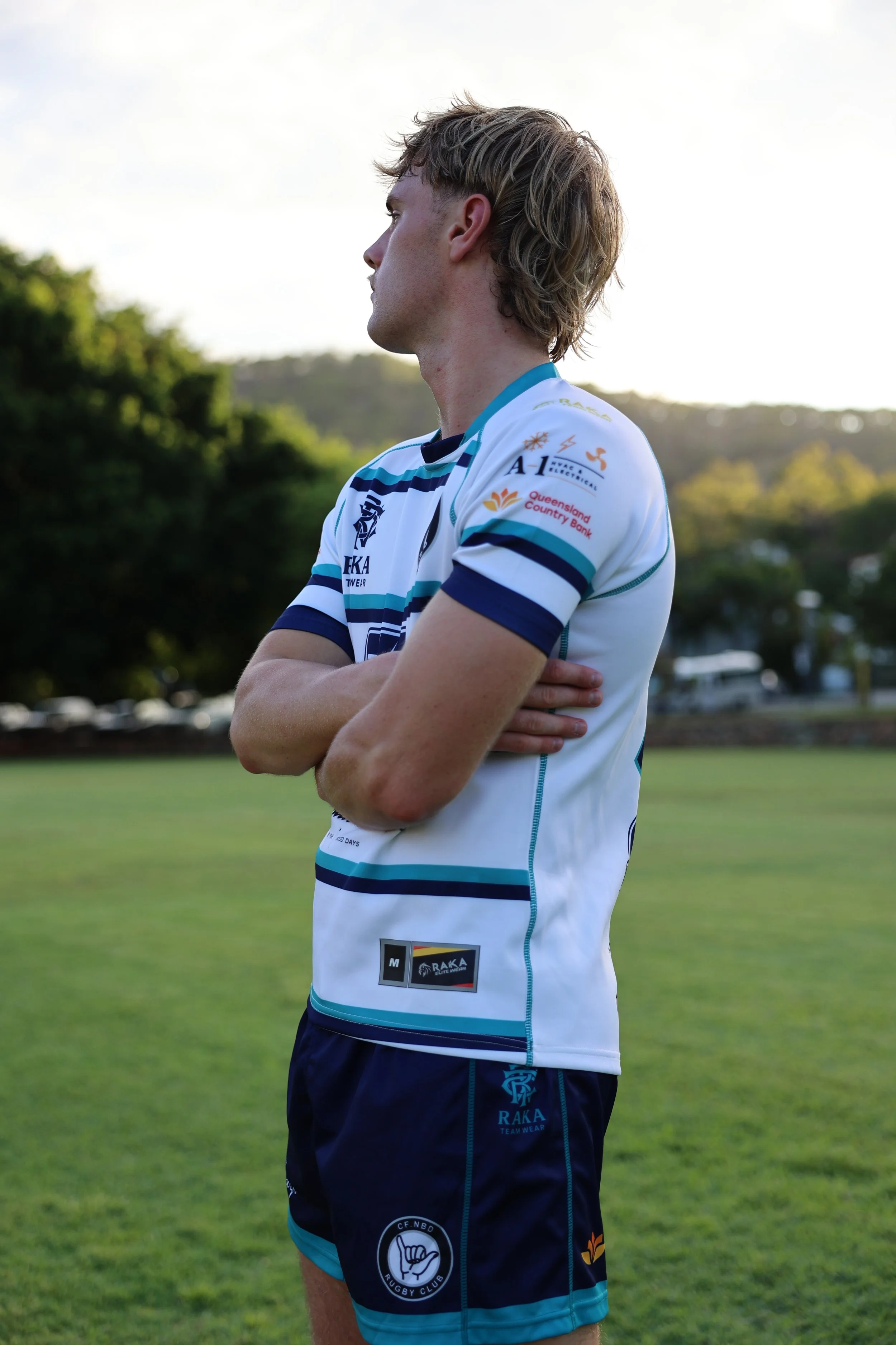 A young man with sunlit blonde hair stands on a grassy field, wearing a white and blue rugby uniform with various sponsor logos, looking contemplative with arms crossed.