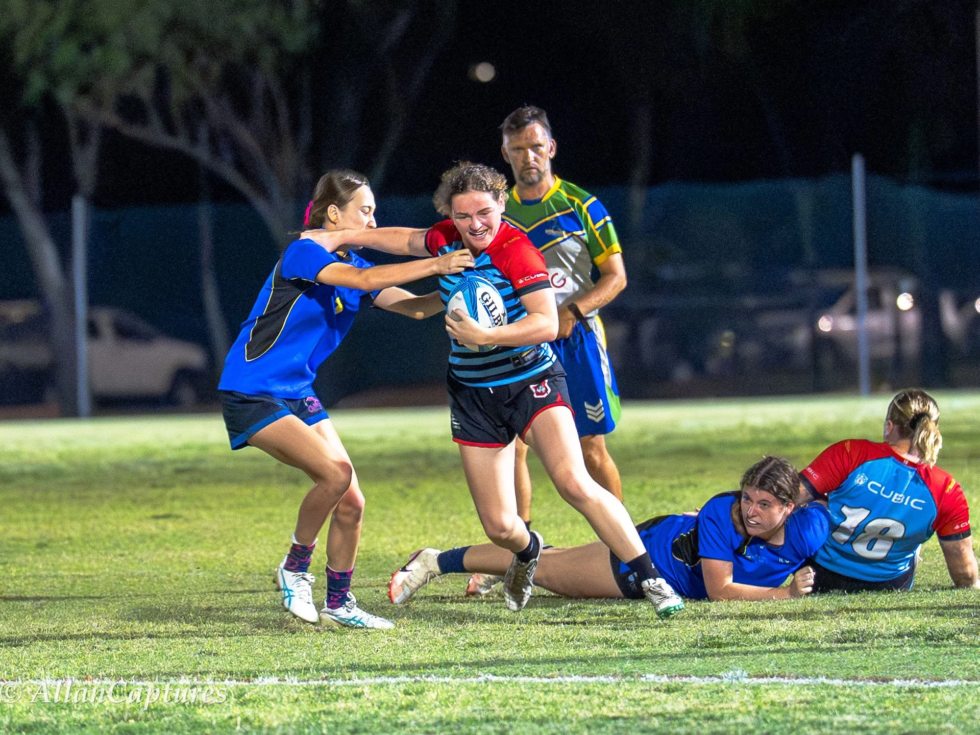 Women playing rugby on a grass field at night, with one player holding a rugby ball and being tackled by another player, while a teammate on the ground and a referee observe.